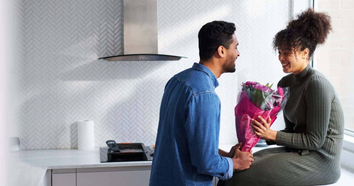 relationship love 桌布 壁紙 — 一名男子向一位坐在現代廚房窗邊微笑的女子獻上一束鮮豔的粉紅色花束。
