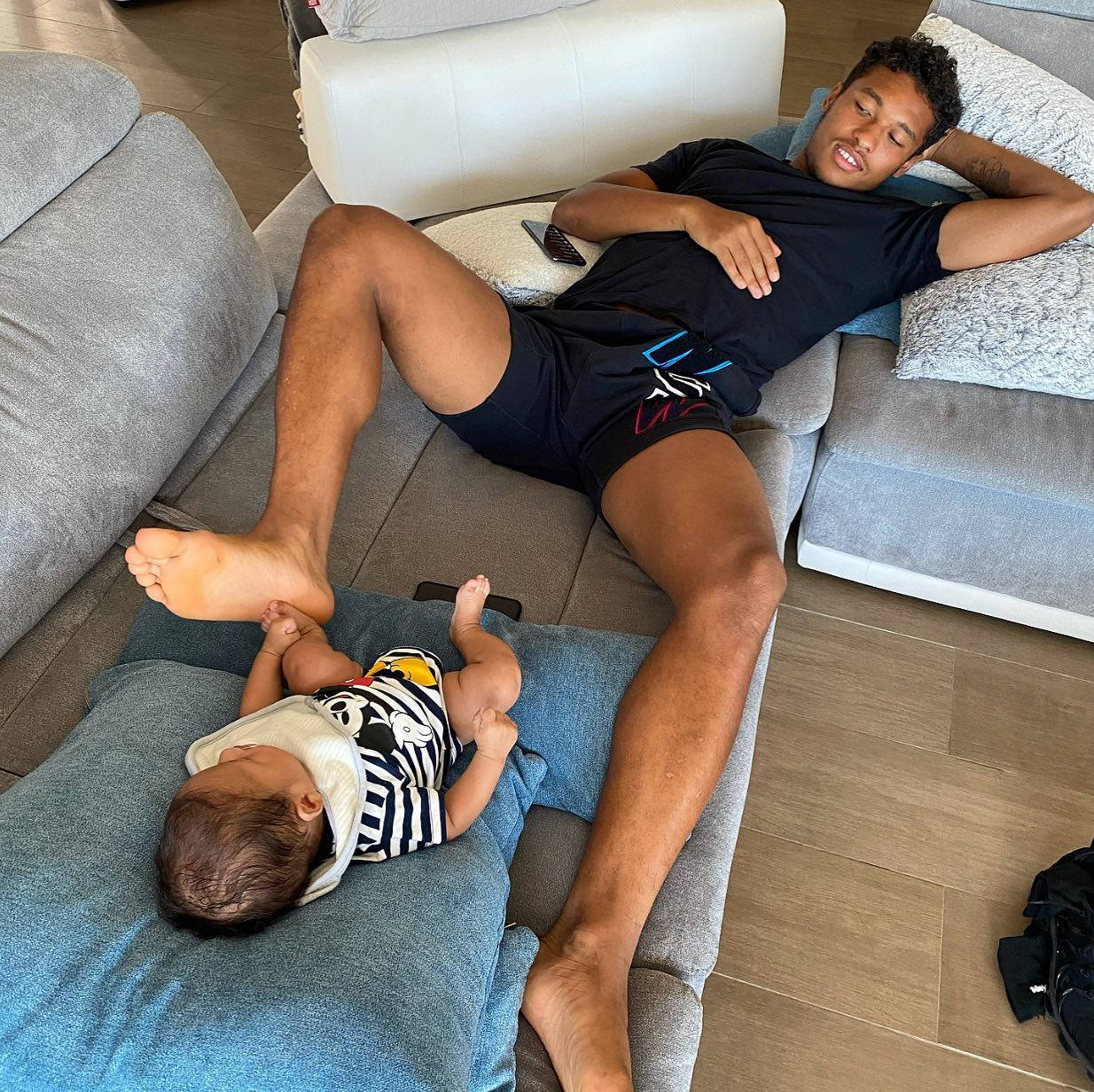 Football Players 桌布 壁紙 — 一名男子和一個嬰兒在舒適的客廳中的現代灰色沙發上平靜地休息。