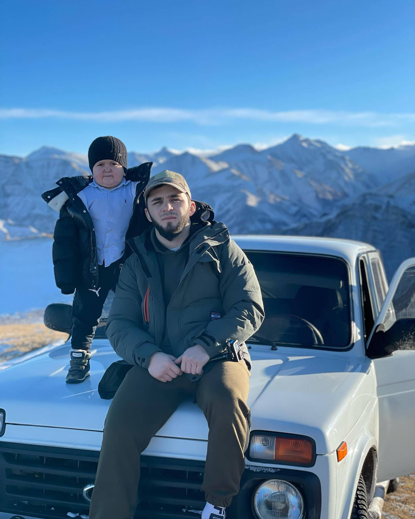 山景 桌布 壁紙 — 一名男子和一個小孩在晴朗藍天下，以積雪山脈為背景，在一輛白色復古車的引擎蓋上擺姿勢。