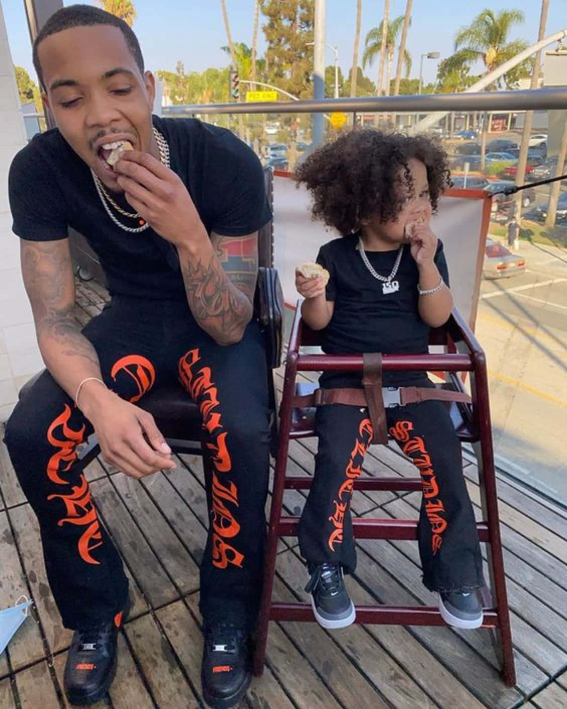Herbo iPhone 桌布 壁紙 — 一名男子和一個小孩穿著相配的黑色和橙色衣服，在一個可以俯瞰陽光普照、種有棕櫚樹的街道的木製甲板上一起吃零食。