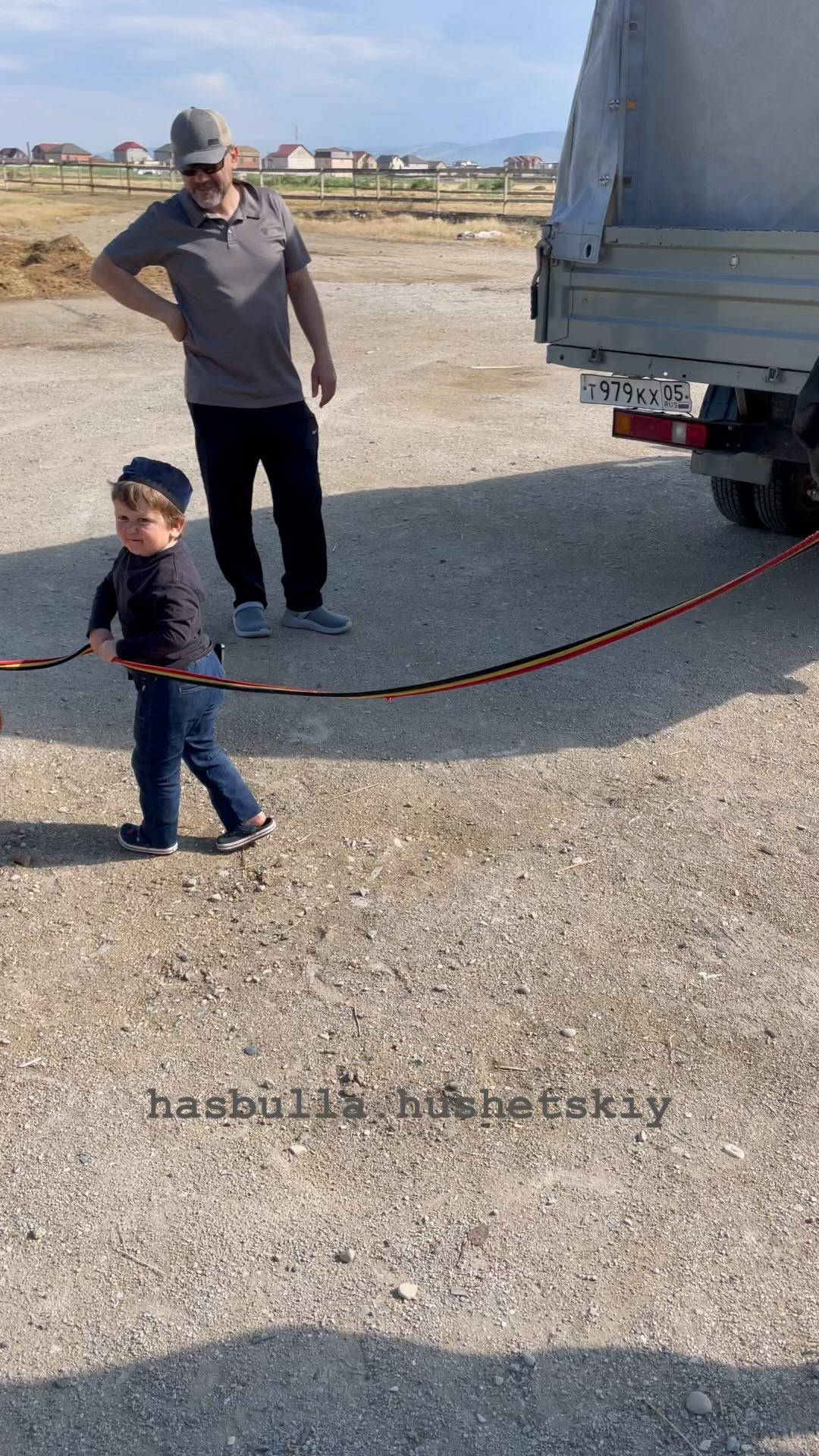 iPhone 桌布 壁紙 — 一名男子和一個幼兒站在碎石上，靠近一輛卡車，他們之間拉著一條紅繩，在明亮的日光下。