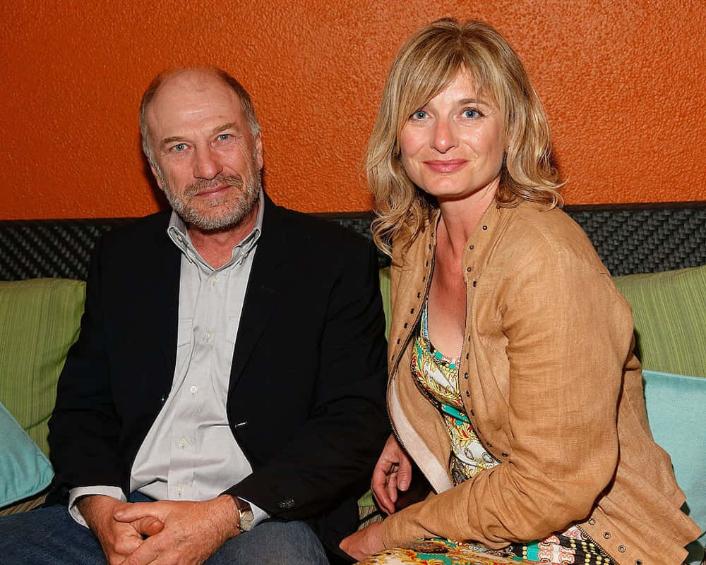 Ted Levine 桌布 壁紙 — 一名男子和一名女子並排坐在沙發上，背景是帶有紋理的橙色牆壁，並有綠色和藍色靠墊。