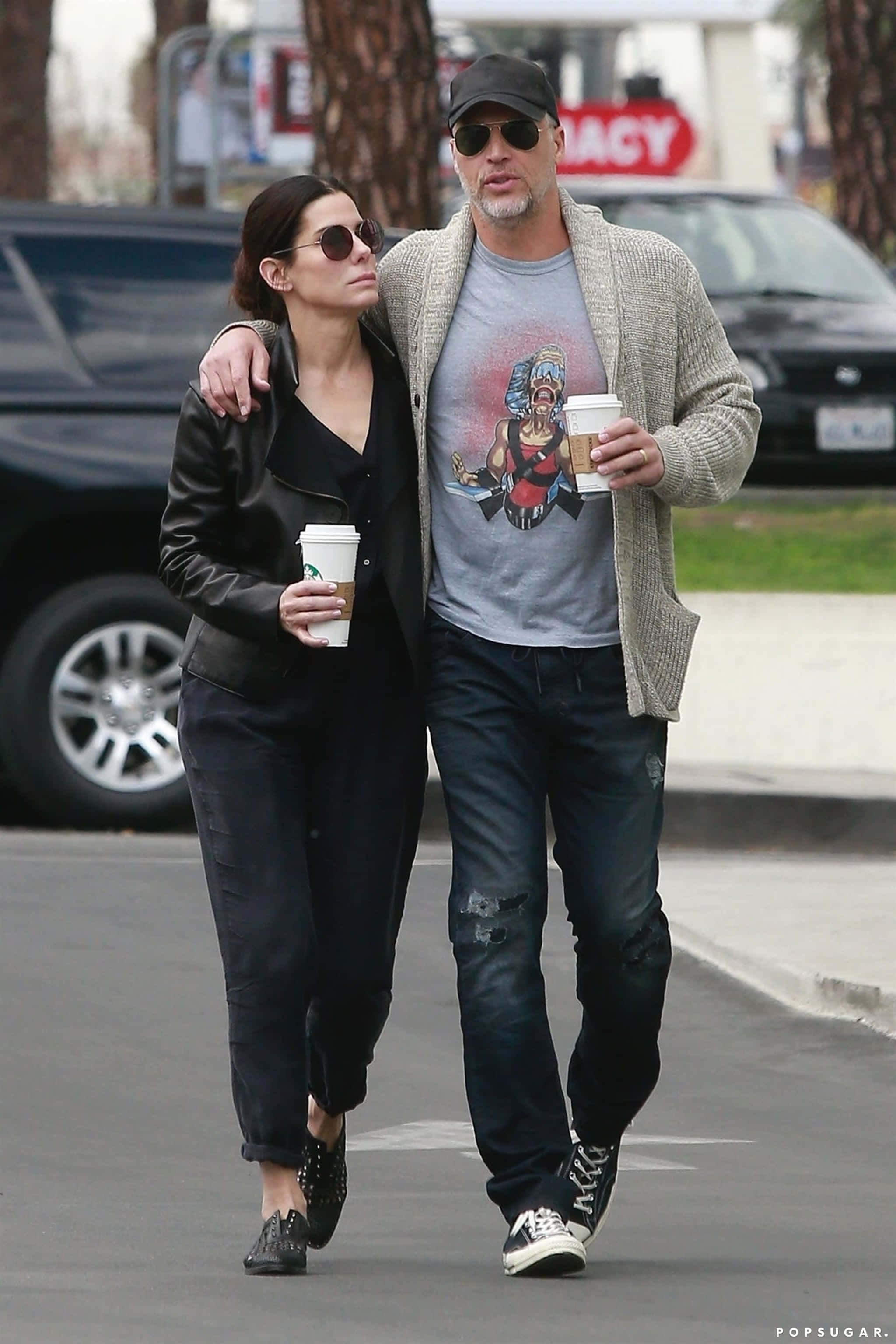 Sandra Bullock 2021 桌布 壁紙 — 一名男子和一名女子各自手持咖啡杯，穿著休閒街頭服飾，悠閒地穿過停車場。