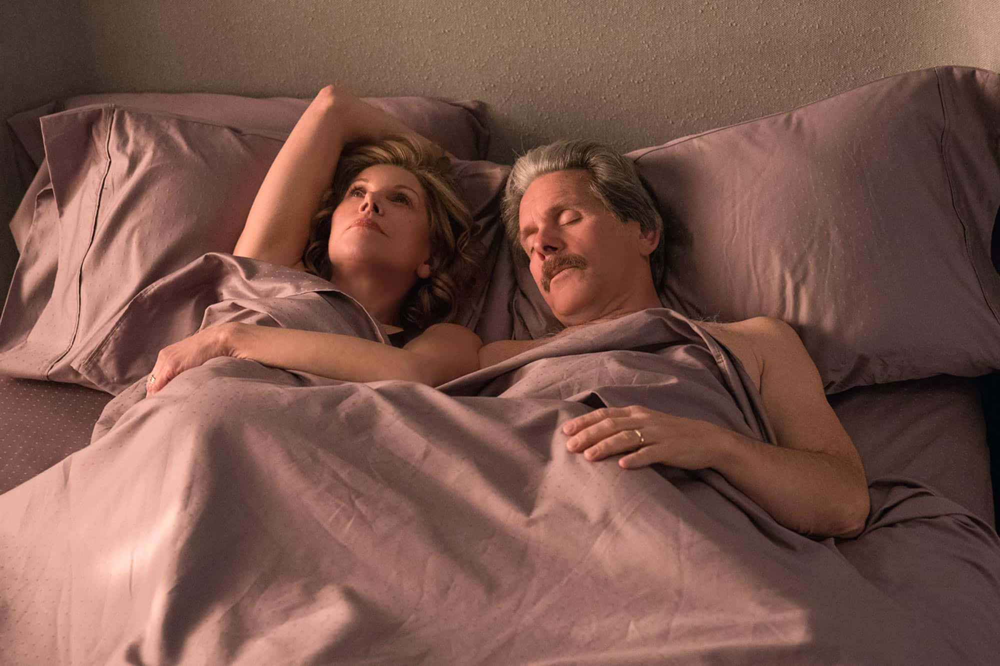 Gary Cole 桌布 壁紙 — 一名男子和一名女子在柔軟的紫色床單下休息，女子醒著向上凝視，而男子則在她旁邊安詳地睡著。