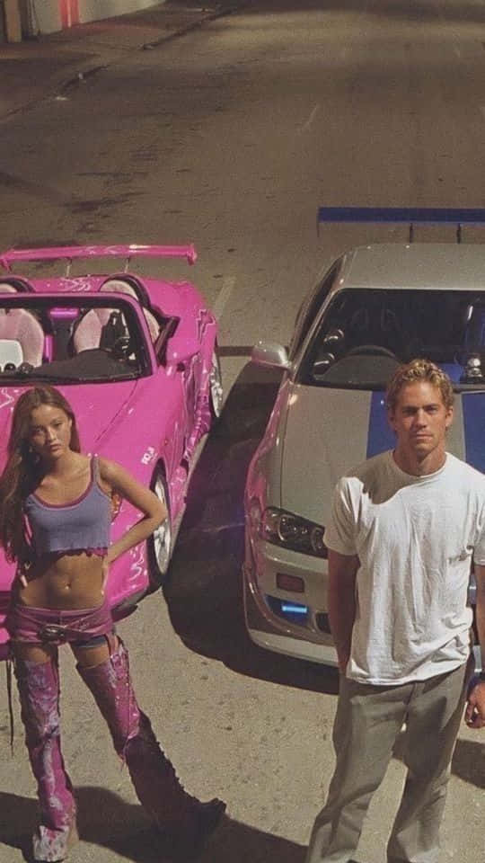 Fast And Furious Desktop 桌布 壁紙 — 一名男子和一名女子站在一輛粉紅色跑車和一輛銀色賽車旁，在夜晚陽光普照的城市街道上。