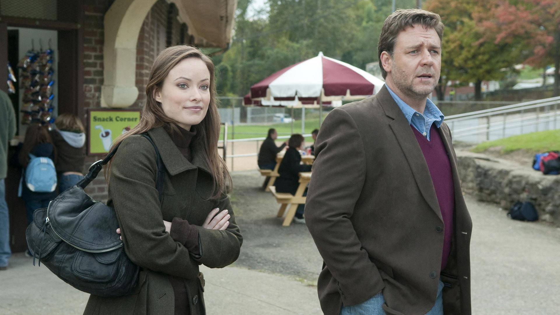 Russell Crowe 桌布 壁紙 — 一名男子和一名女子站在學校外面，雙臂交叉，表情嚴肅，學生們則坐在附近的野餐桌旁。