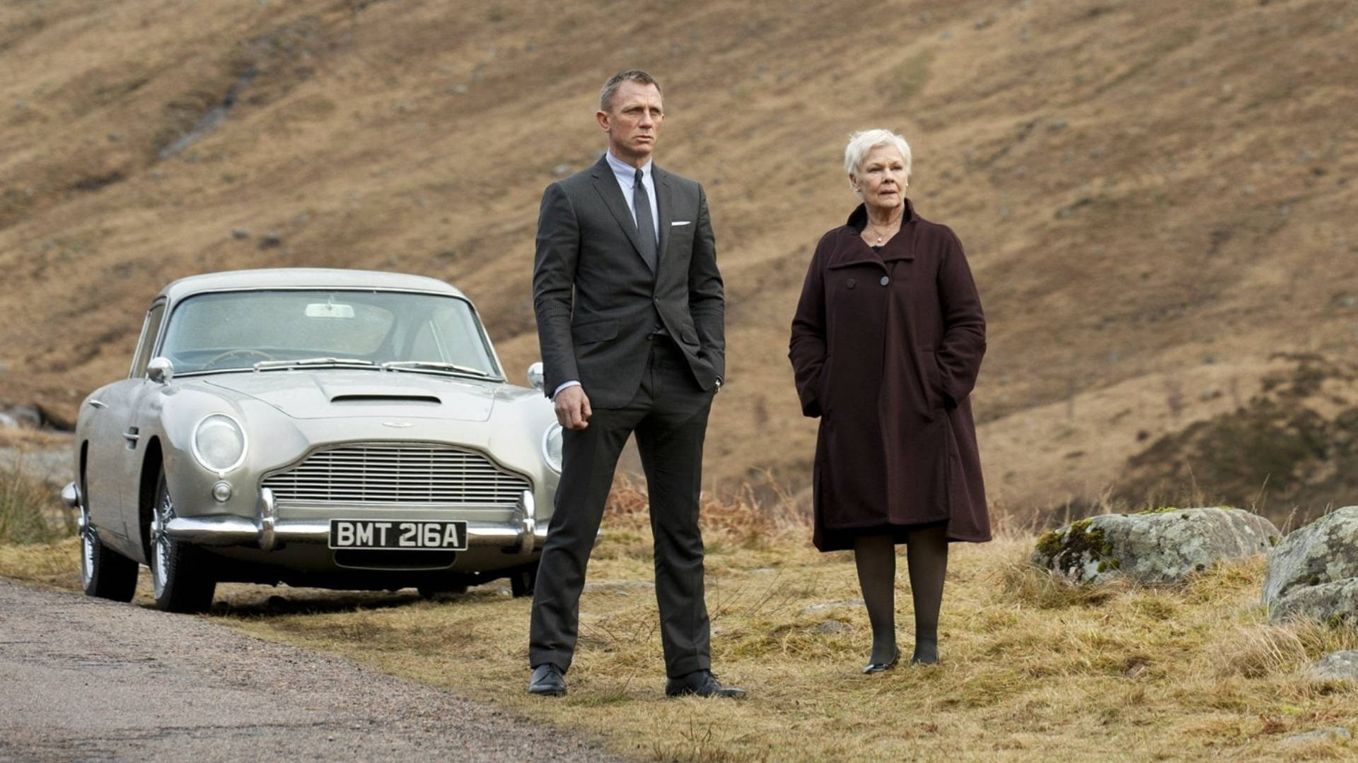 Daniel Craig 桌布 壁紙 — 一名男子和一名女子站在碎石路上，旁邊是一輛銀色 Aston Martin DB5，背景是乾燥的草坡。