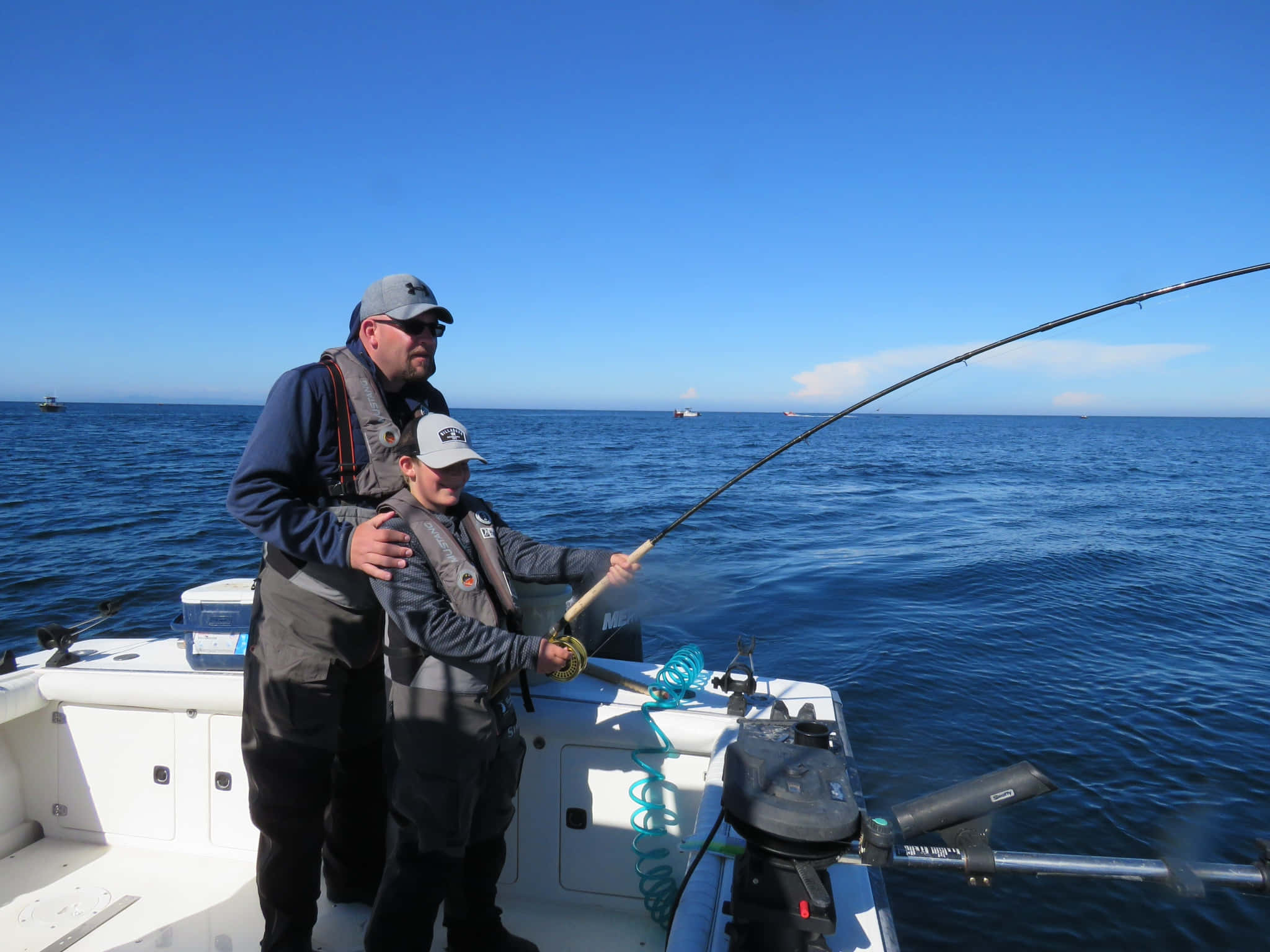 Fishing Rod 桌布 壁紙 — 一名男子和一名男孩穿著救生衣，在晴朗藍天下平靜的海面上，於白色船上一起釣魚。