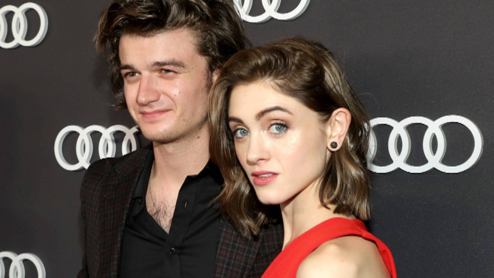 Joe Keery 桌布 壁紙 — 一名男子和女子在奧迪品牌活動中合照，背景為深色，帶有重複的白色標誌。