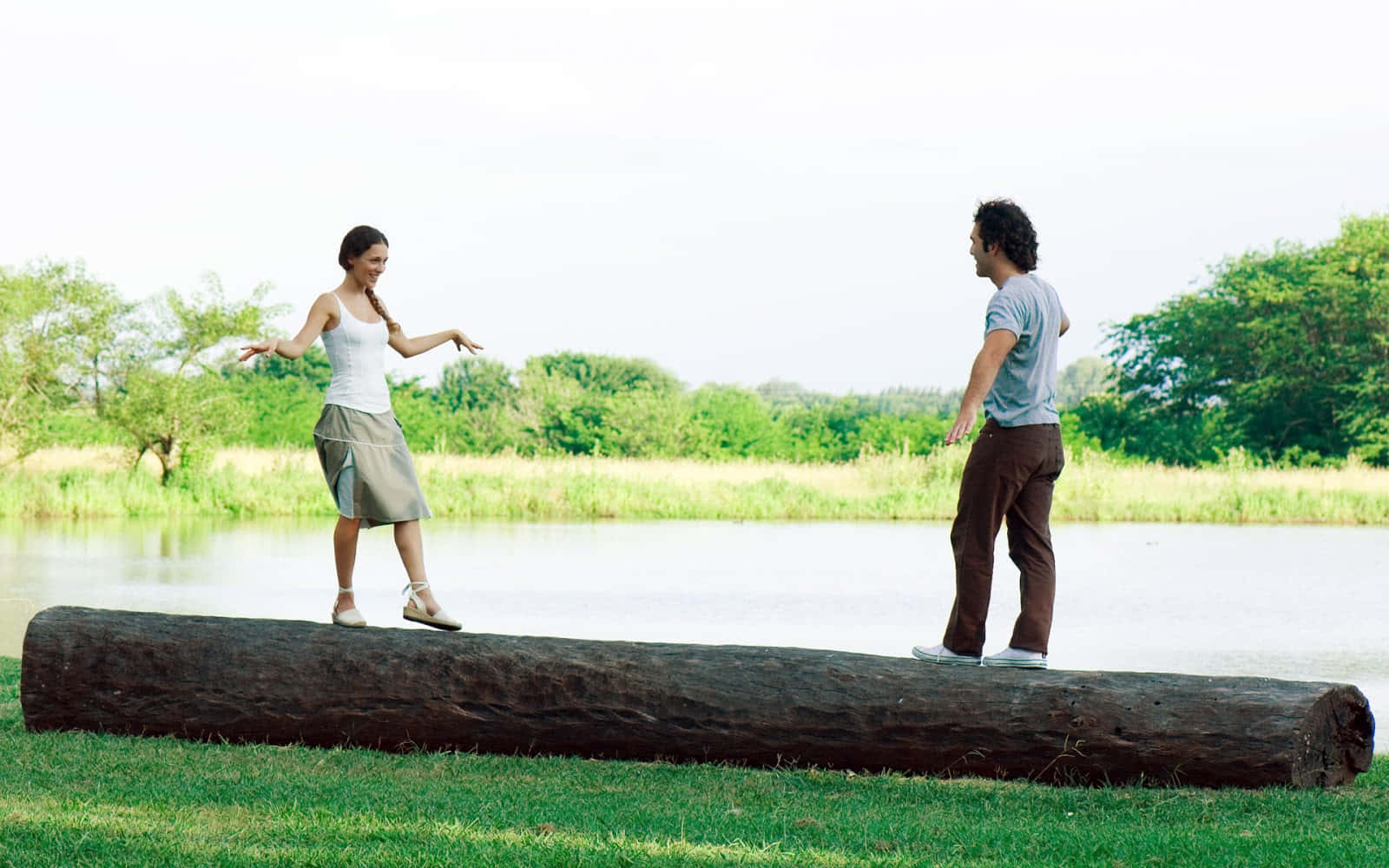 Lake 桌布 壁紙 — 一名男子和女子在平靜的湖邊，在茂密綠樹環繞下，於明亮的日光中在一根大木頭上保持平衡。