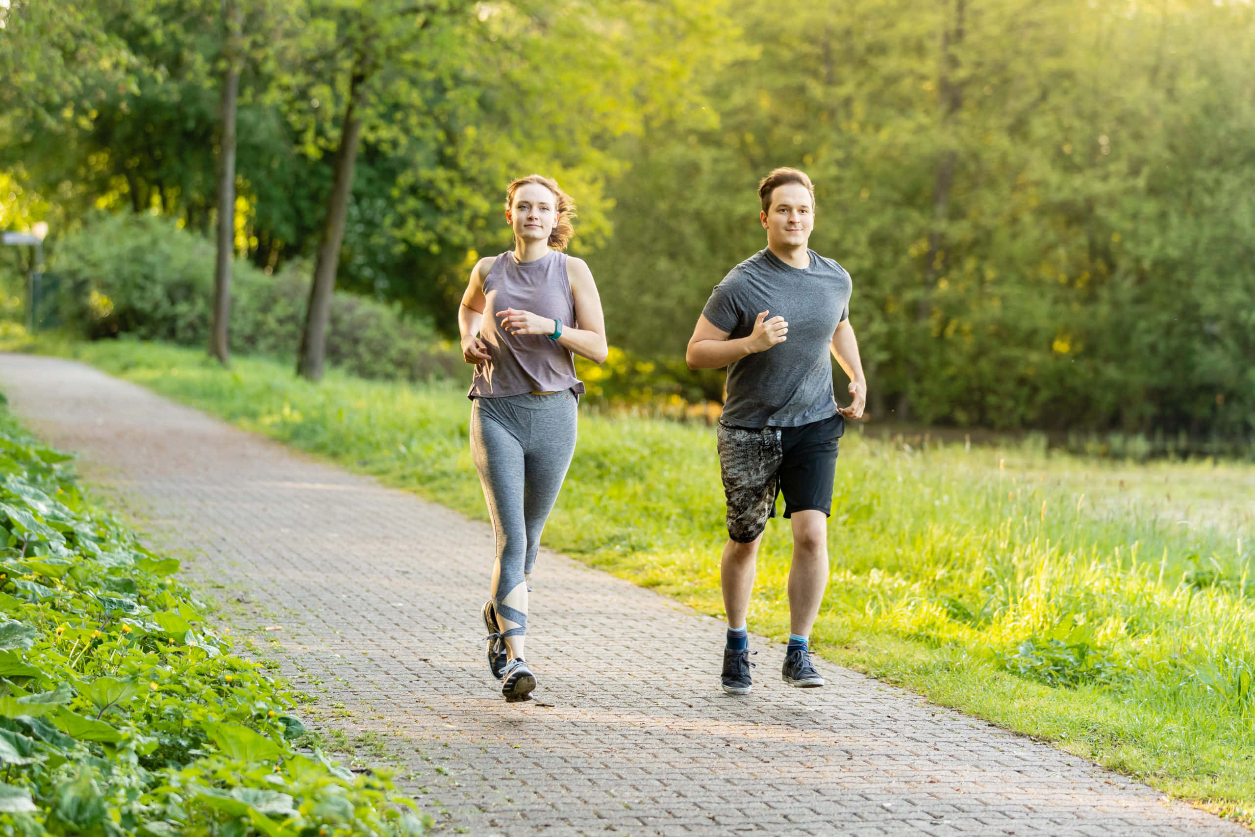 Exercise 桌布 壁紙 — 一名男子和女子在陽光普照的磚徑上並肩慢跑，穿過翠綠茂盛的公園。