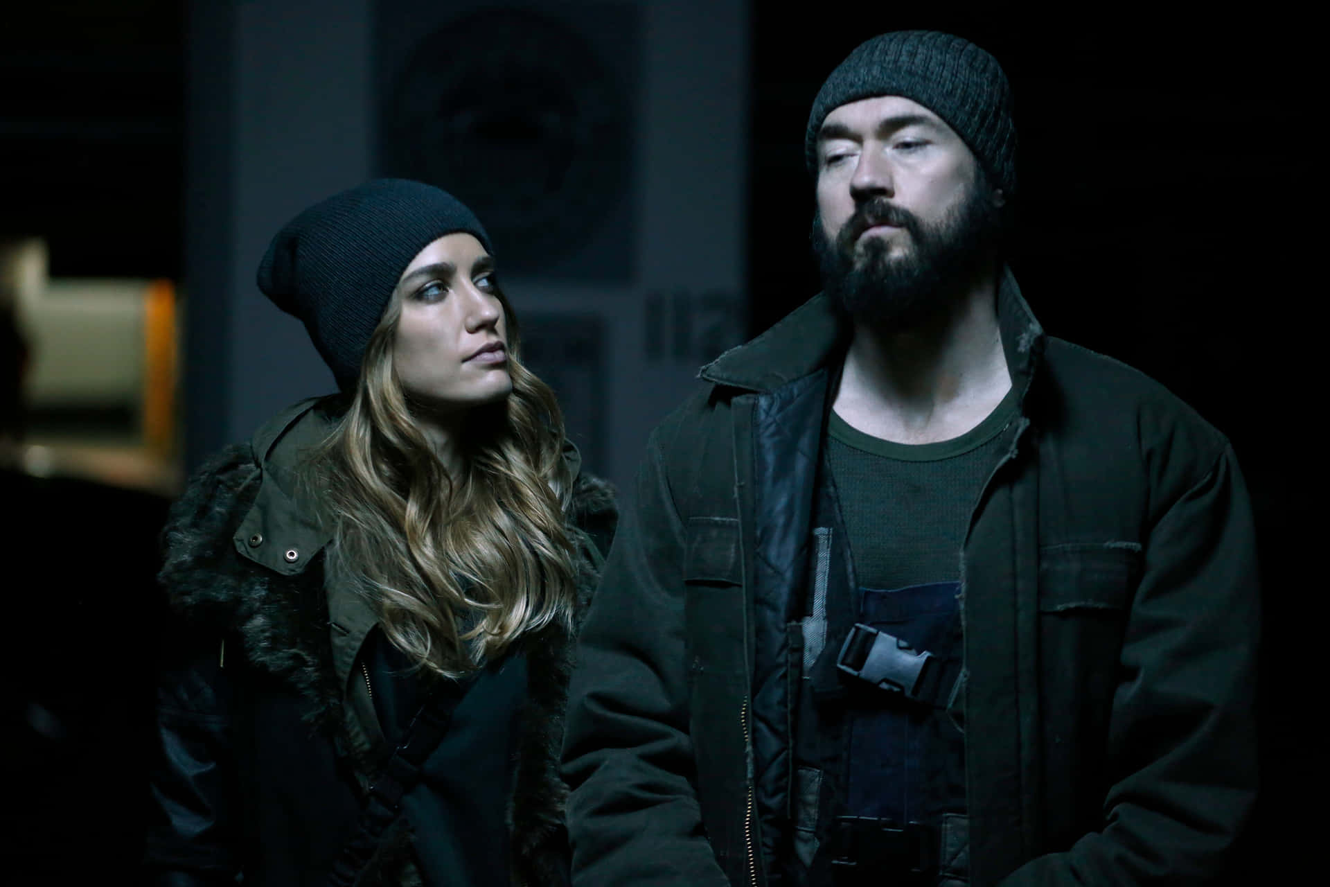Kevin Durand 桌布 壁紙 — 一名男子和女子穿著深色冬季裝備，站在燈光昏暗的房間裡，表情嚴肅。