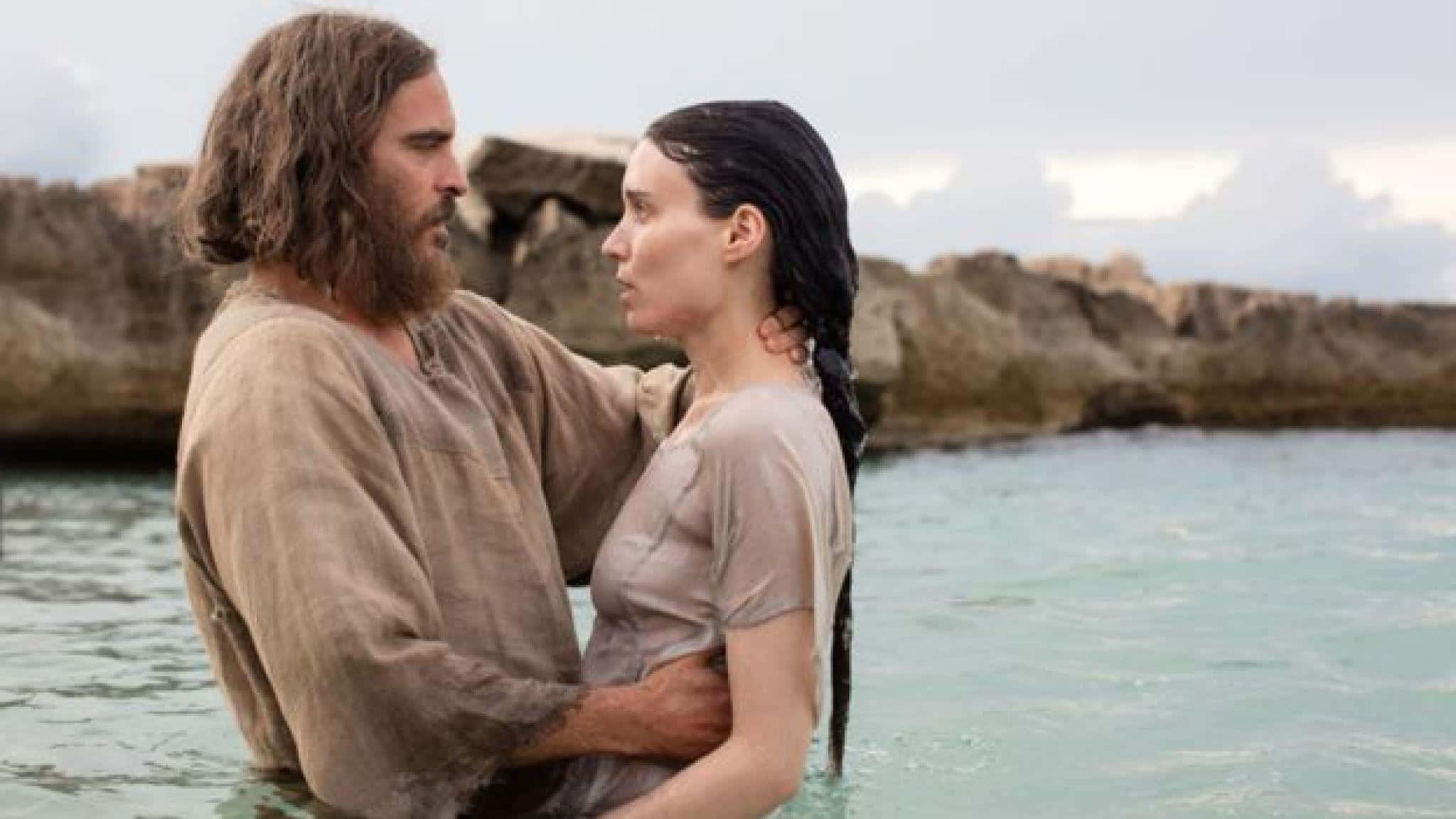 Mary Magdalene 桌布 壁紙 — 一名男子和女子站在平靜的藍綠色水中，水深及腰，在多雲的天空下以崎嶇的懸崖為背景擁抱。