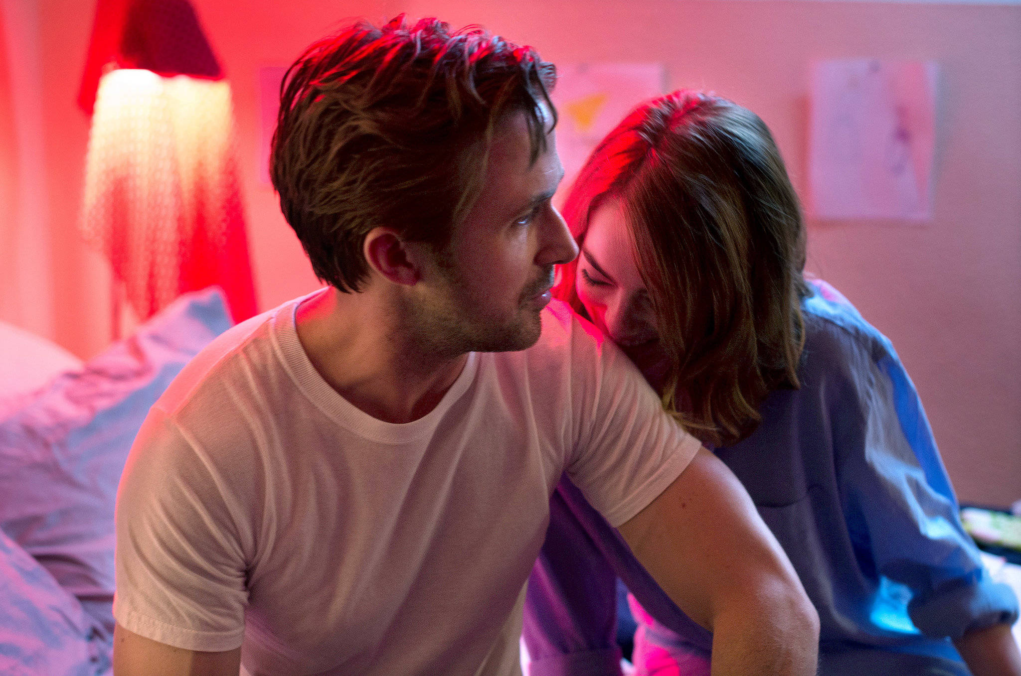 Ryan Gosling 桌布 壁紙 — 一名男子和女子親密地坐在床上，溫暖的紅燈從燈具灑落，在他們身後的牆上投下柔和的陰影。