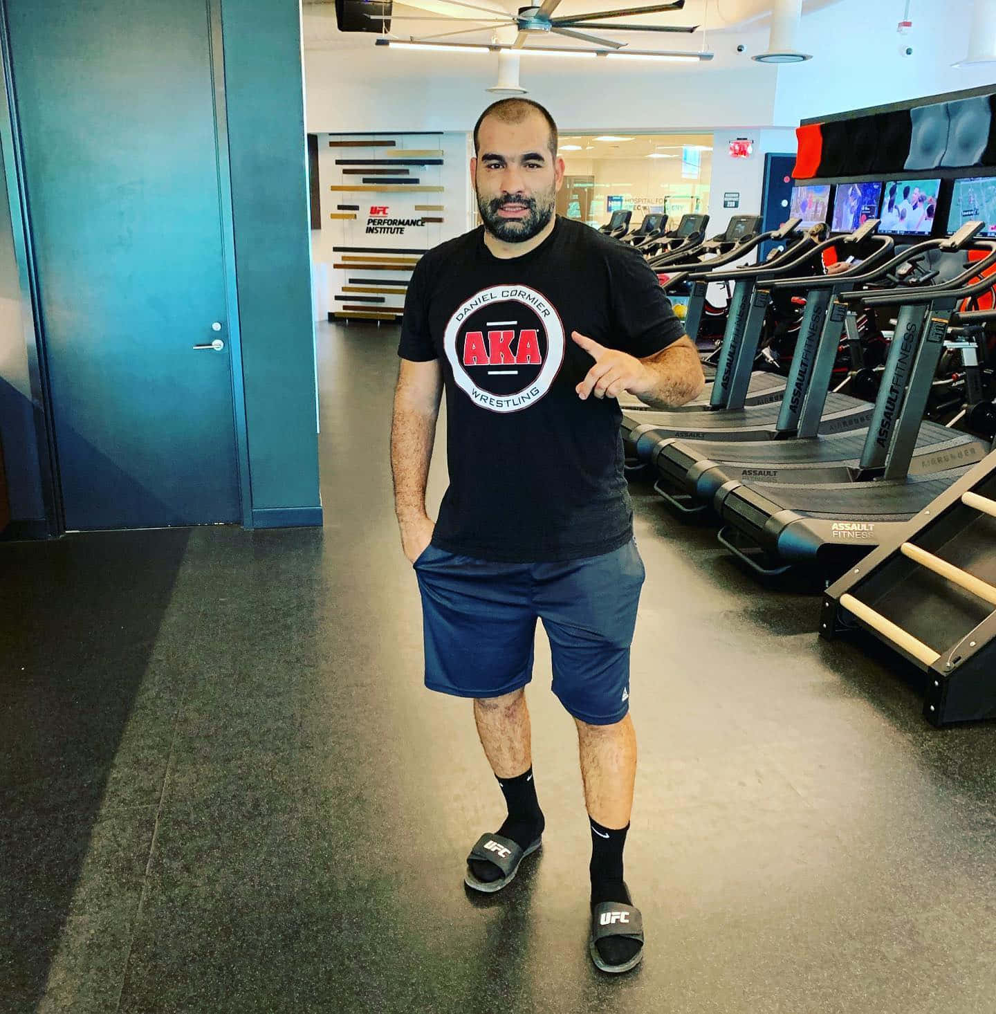 健身房 桌布 壁紙 — 一名男子在UFC健身房自豪地指著他的AKA Wrestling T恤，站在跑步機附近。