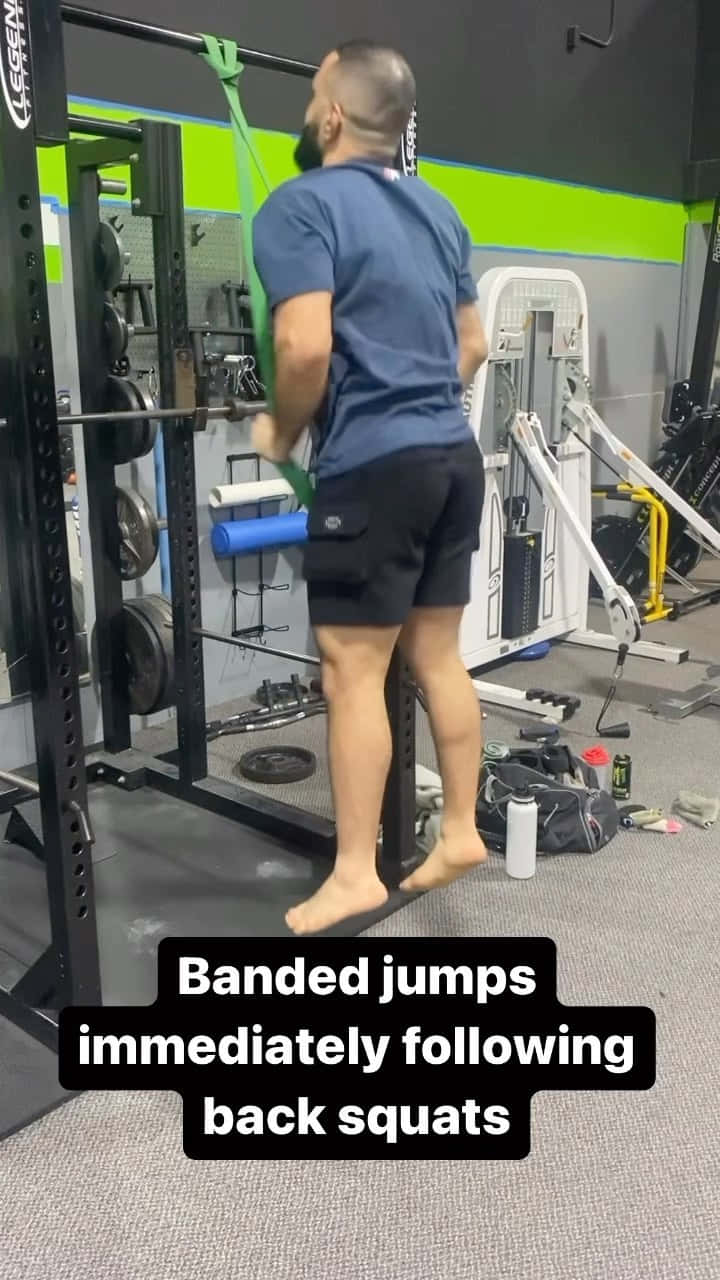 Fitness 桌布 壁紙 — 一名男子在健身房赤腳進行彈力帶跳躍，附近有深蹲架，周圍有槓鈴和器材。