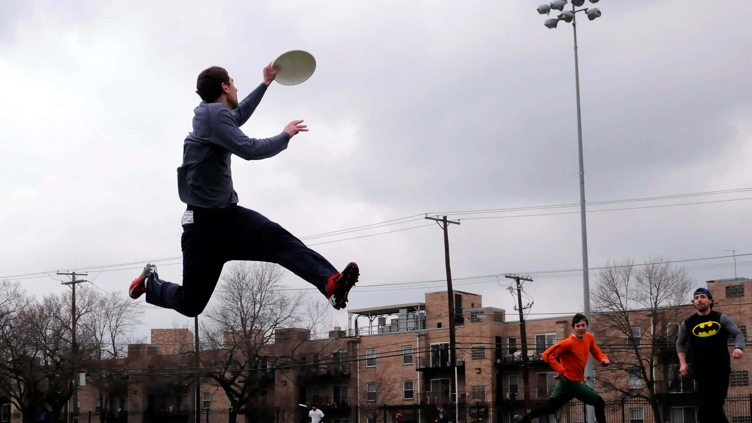 1440p Ultimate Frisbee 桌布 壁紙 — 一名男子在城市公園中躍起接住飛盤，另外兩人在陰天天空下觀看。