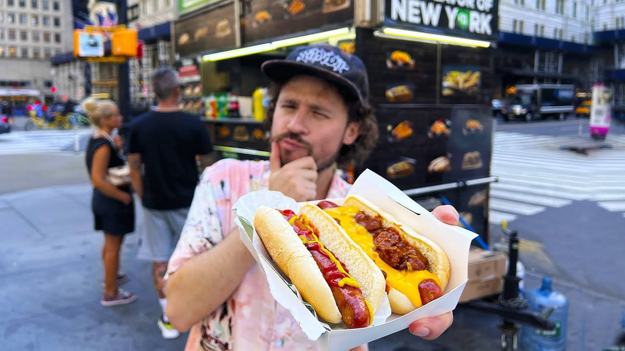 Hot Dog 桌布 壁紙 — 一名男子在城市的街道上，手持兩份加了芥末、番茄醬和辣椒的熱狗，神情若有所思。