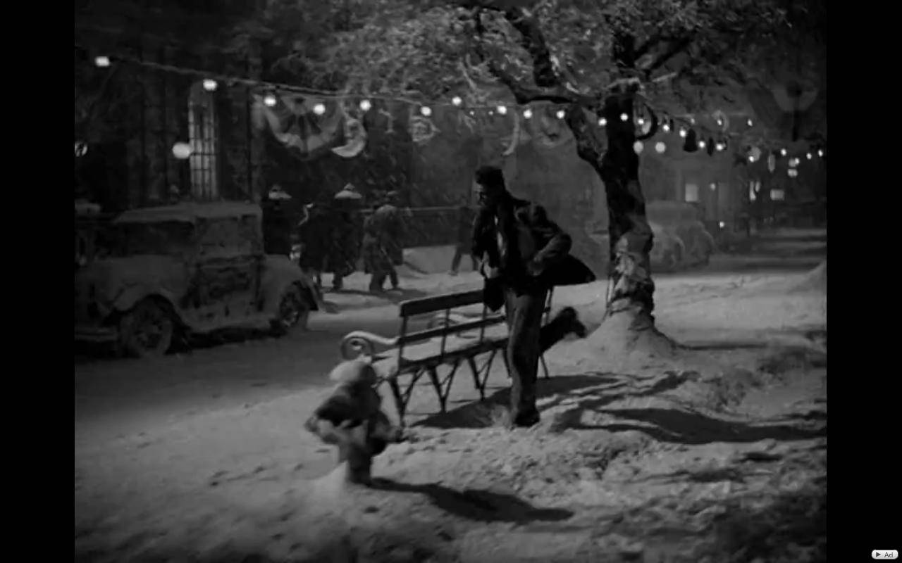 Film 桌布 壁紙 — 一名男子在夜晚的雪街上，在燈串下推著一張被雪覆蓋的長椅，背景有復古汽車和行人。