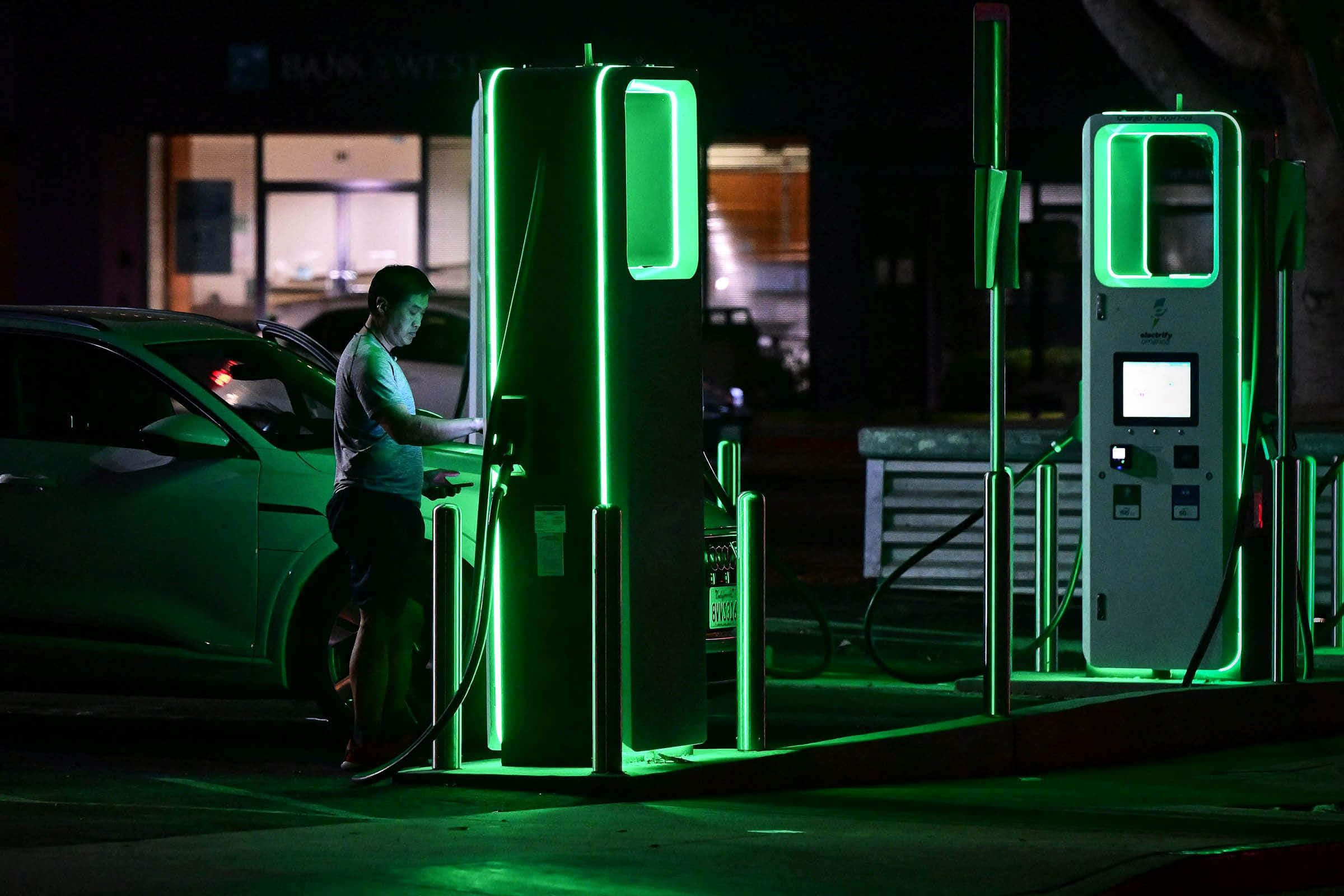 Electric Vehicle 桌布 壁紙 — 一名男子在夜空下將他的電動車插入發光的綠色充電站。