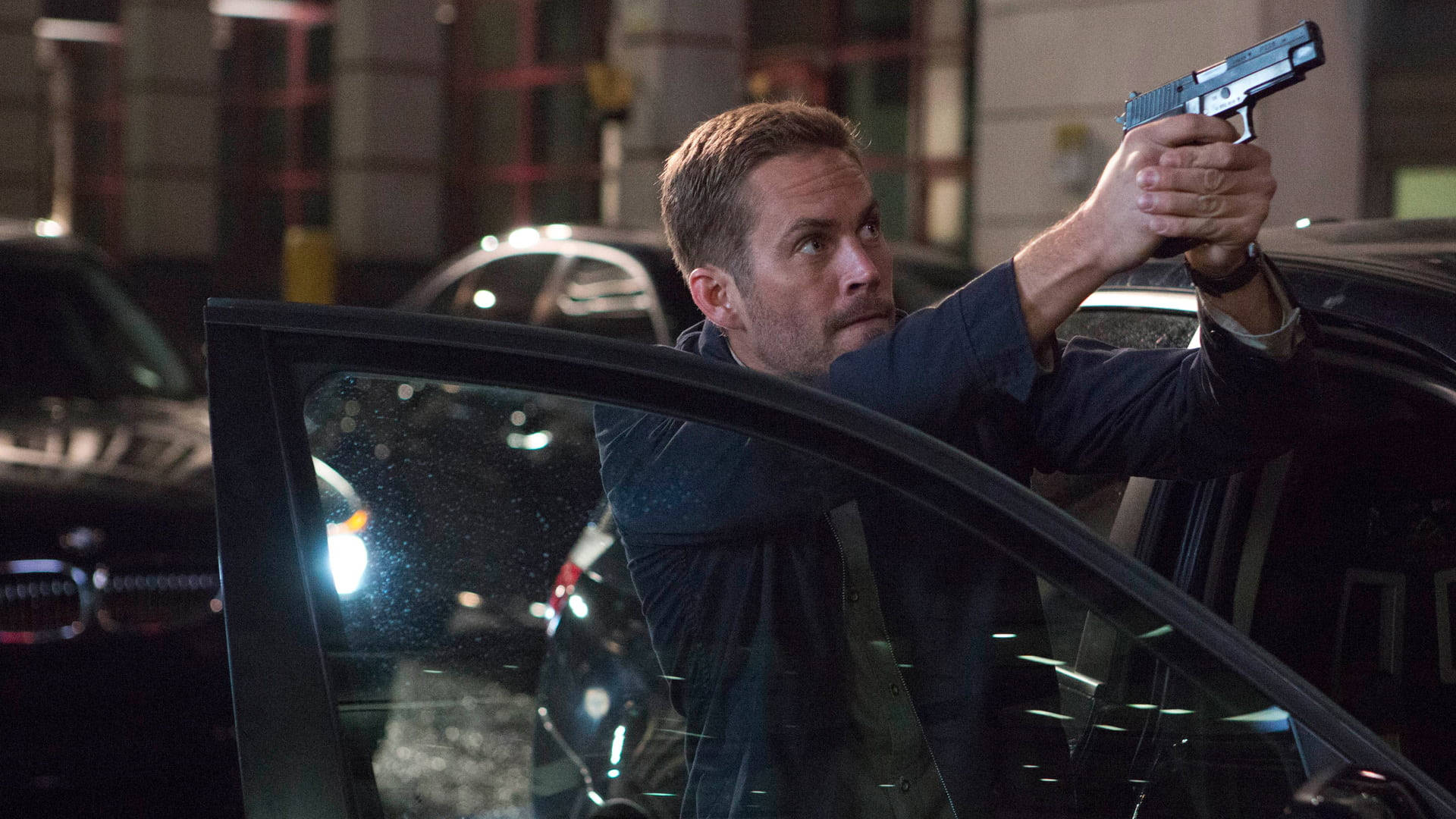 Paul Walker 桌布 壁紙 — 一名男子在夜間從車窗探出身子，專注地瞄準手槍，身處城市環境。