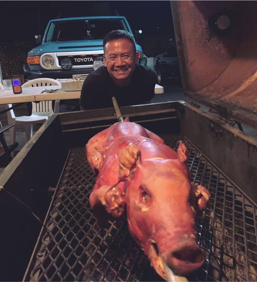 Pig 桌布 壁紙 — 一名男子在夜間自豪地微笑，旁邊是一個烤全豬在烤架上，身後有一輛藍色豐田車。