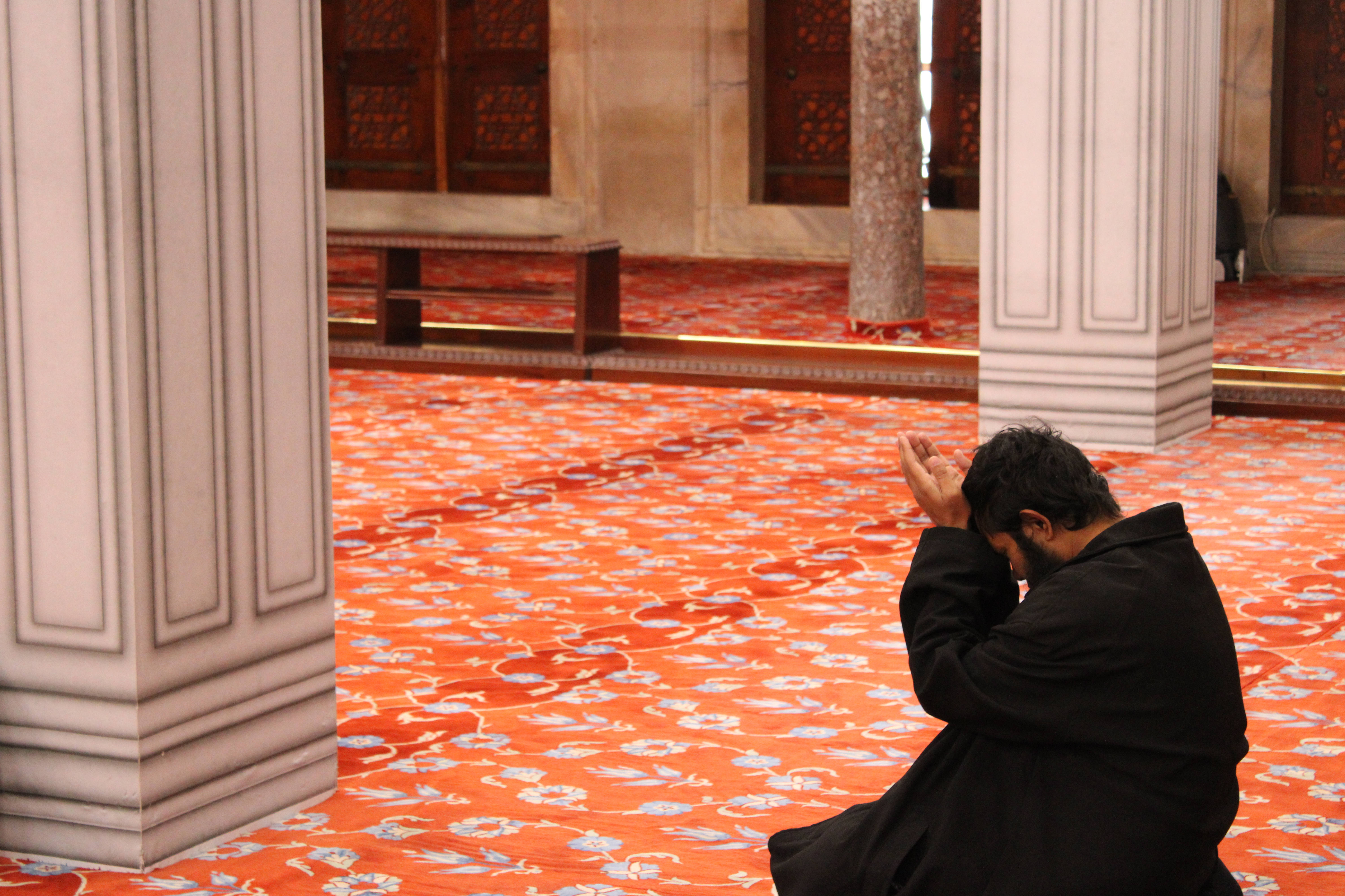 Ramadan Mubarak 桌布 壁紙 — 一名男子在宏偉的清真寺內，在裝飾性的橙色花卉地毯上跪著祈禱，寺內有白色柱子和木門。