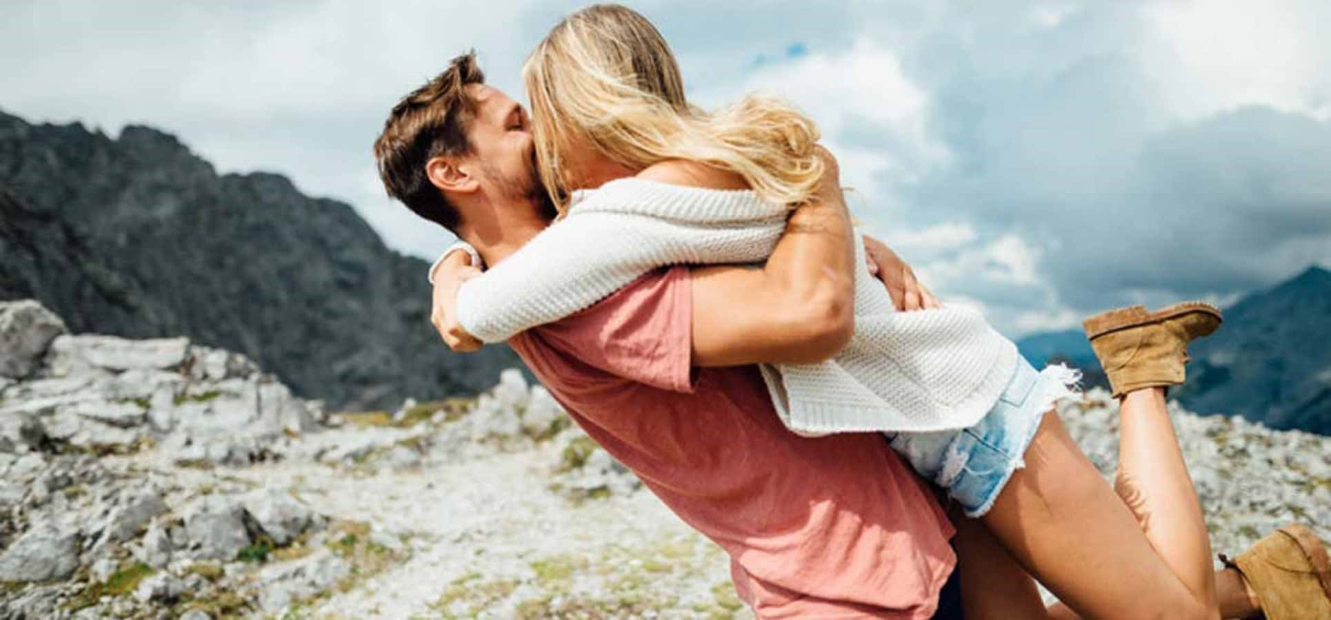 Falling In Love 桌布 壁紙 — 一名男子在崎嶇山脈和陰雲密佈的天空背景下，歡樂地擁抱並舉起一名女子。