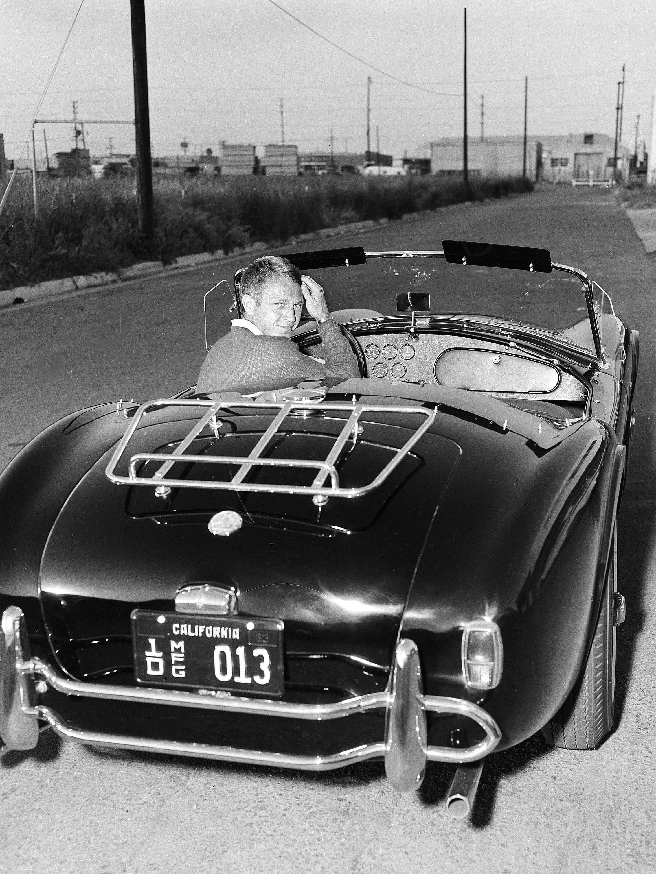 Black And White Vintage Car 桌布 壁紙 — 一名男子在復古黑色敞篷車上微笑，同時在明亮的日光下於安靜的路邊整理頭髮。