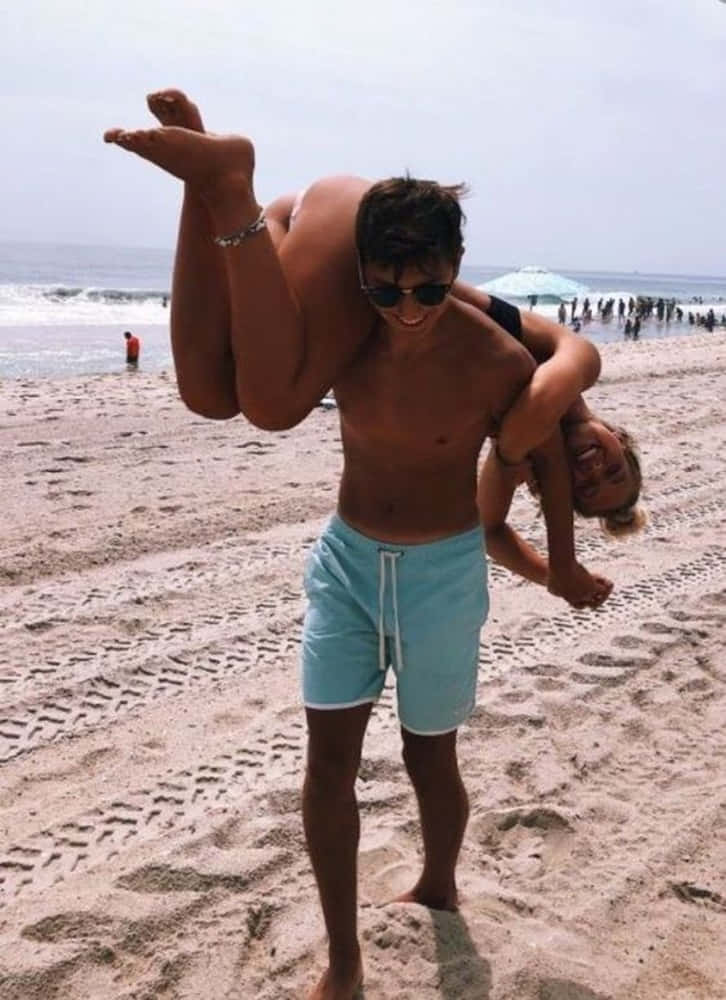 Girlfriend And Boyfriend 桌布 壁紙 — 一名男子在擁擠的海灘上愉快地將一名女子倒轉抱起，背後有海浪和陰天。