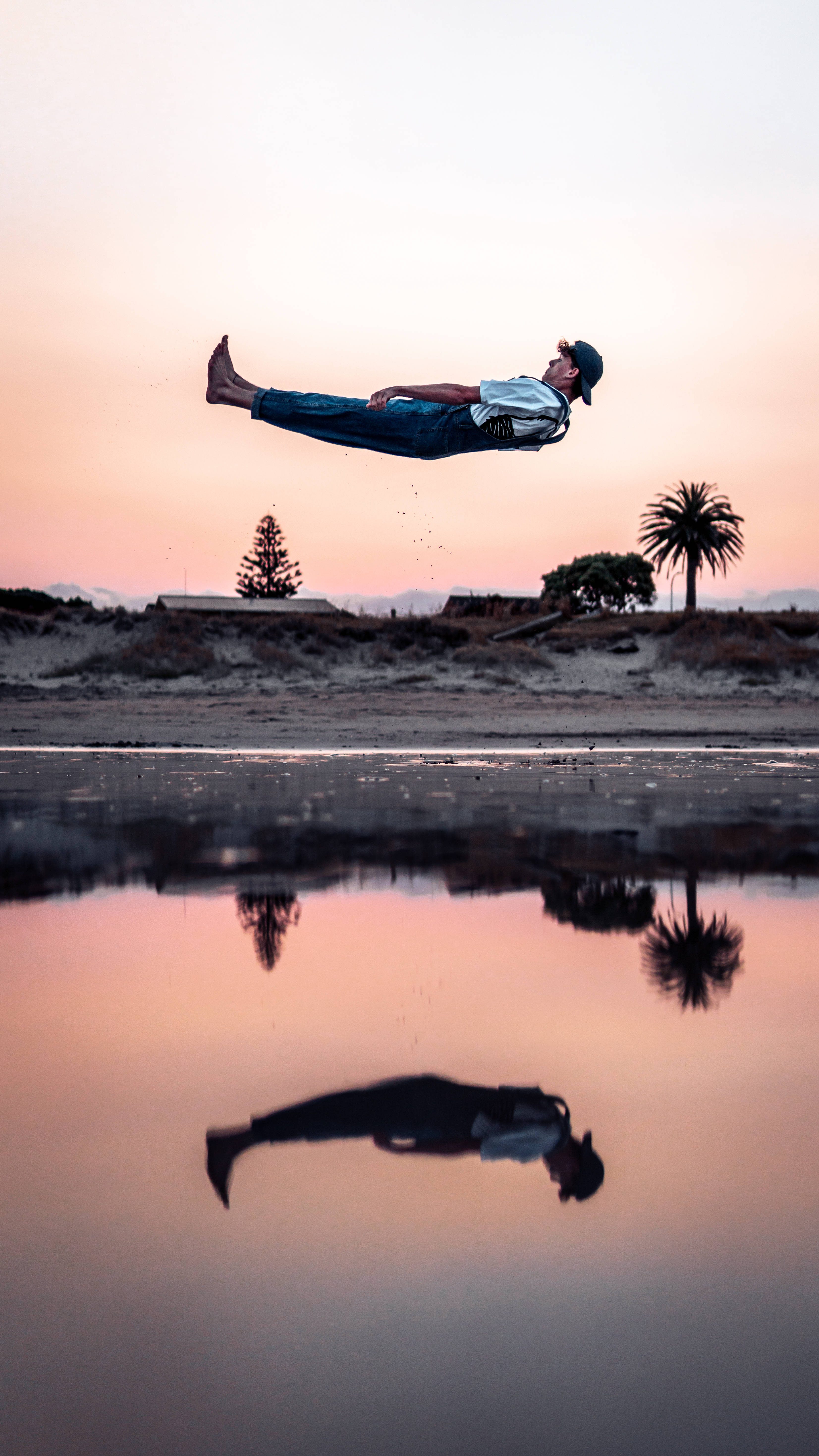 Parkour 桌布 壁紙 — 一名男子在日落時分漂浮在反射水坑上方，背景有棕櫚樹和柔和的粉紅色天空。