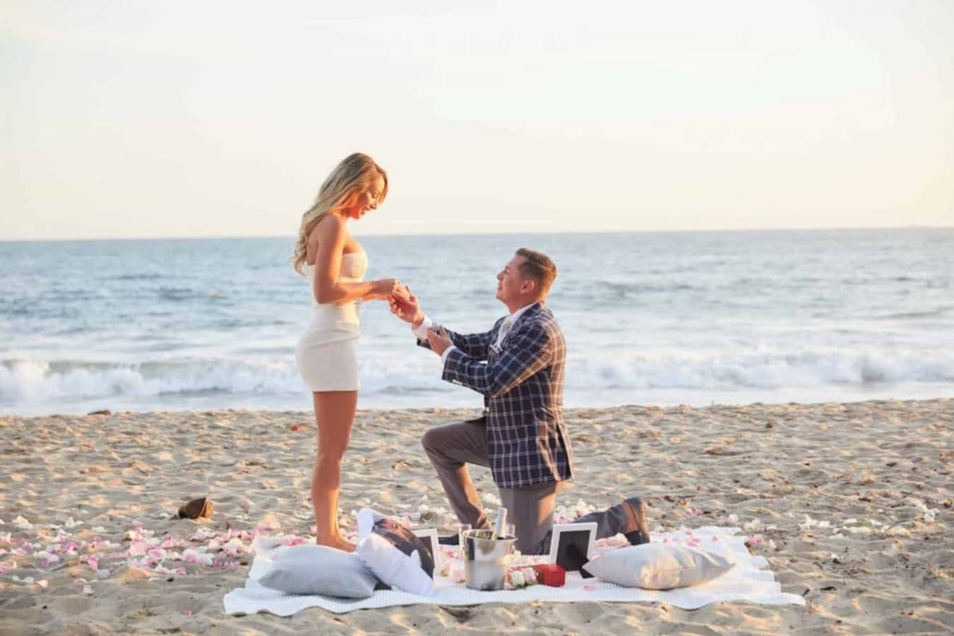 Proposal 桌布 壁紙 — 一名男子在日落時分跪在海灘上，向一位身穿白色連衣裙、被粉紅玫瑰花瓣和香檳包圍的女子求婚。