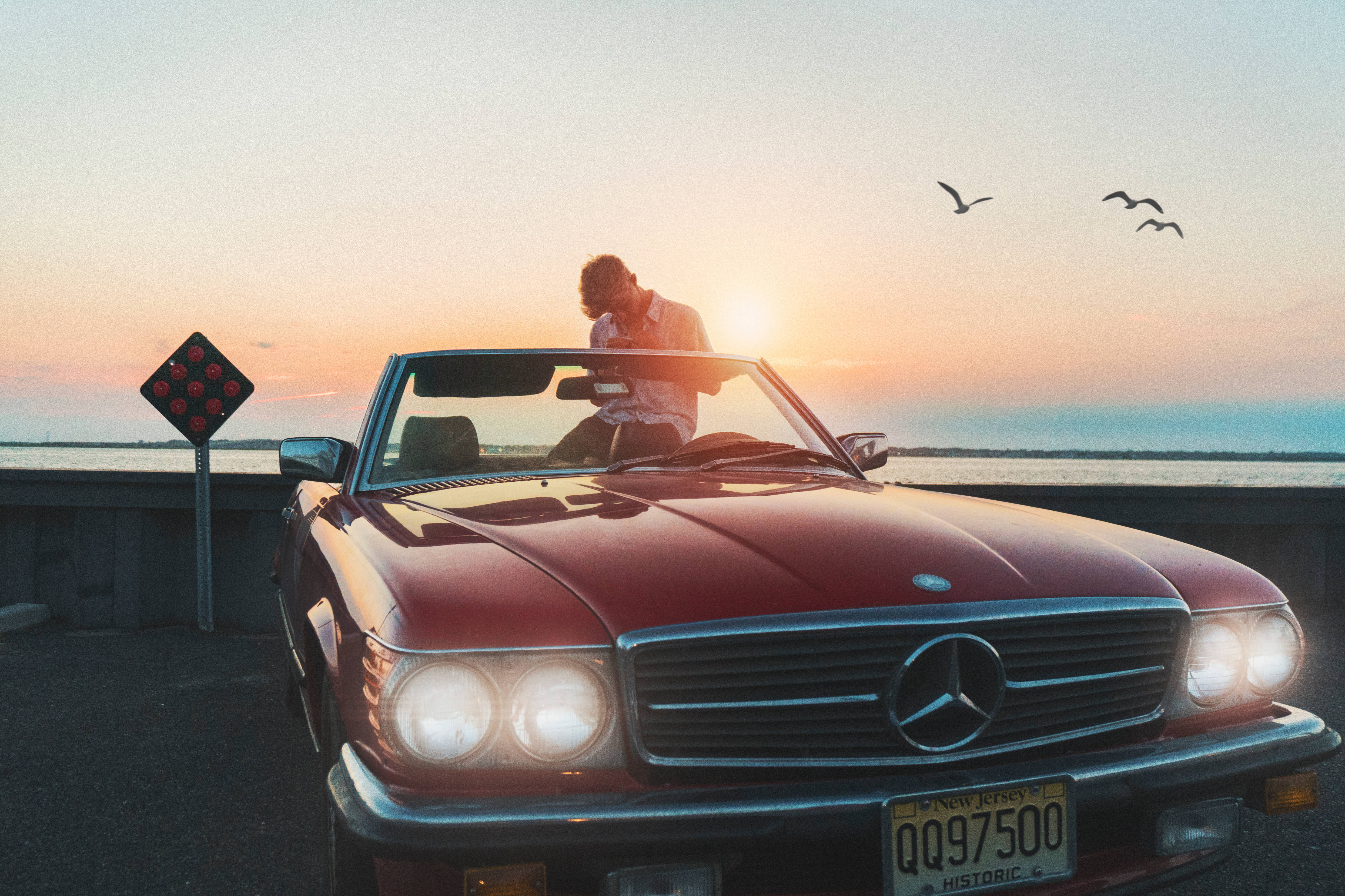 高畫質 賓士 桌布 壁紙 — 一名男子在日落時分靠向一輛紅色敞篷Mercedes-Benz，頭頂有鳥飛過，背景有水。