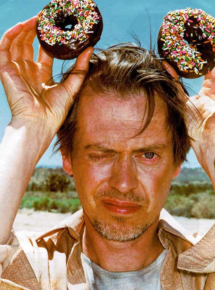steve buscemi 桌布 壁紙 — 一名男子在明亮的藍天和沙漠背景下，將兩個巧克力彩針甜甜圈放在頭上。