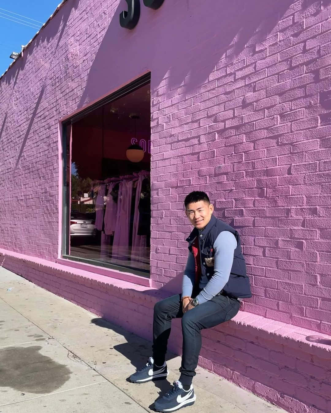 Mens Fashion 桌布 壁紙 — 一名男子在晴朗的藍天下，隨意地坐在商店外亮粉紅色的磚牆上。