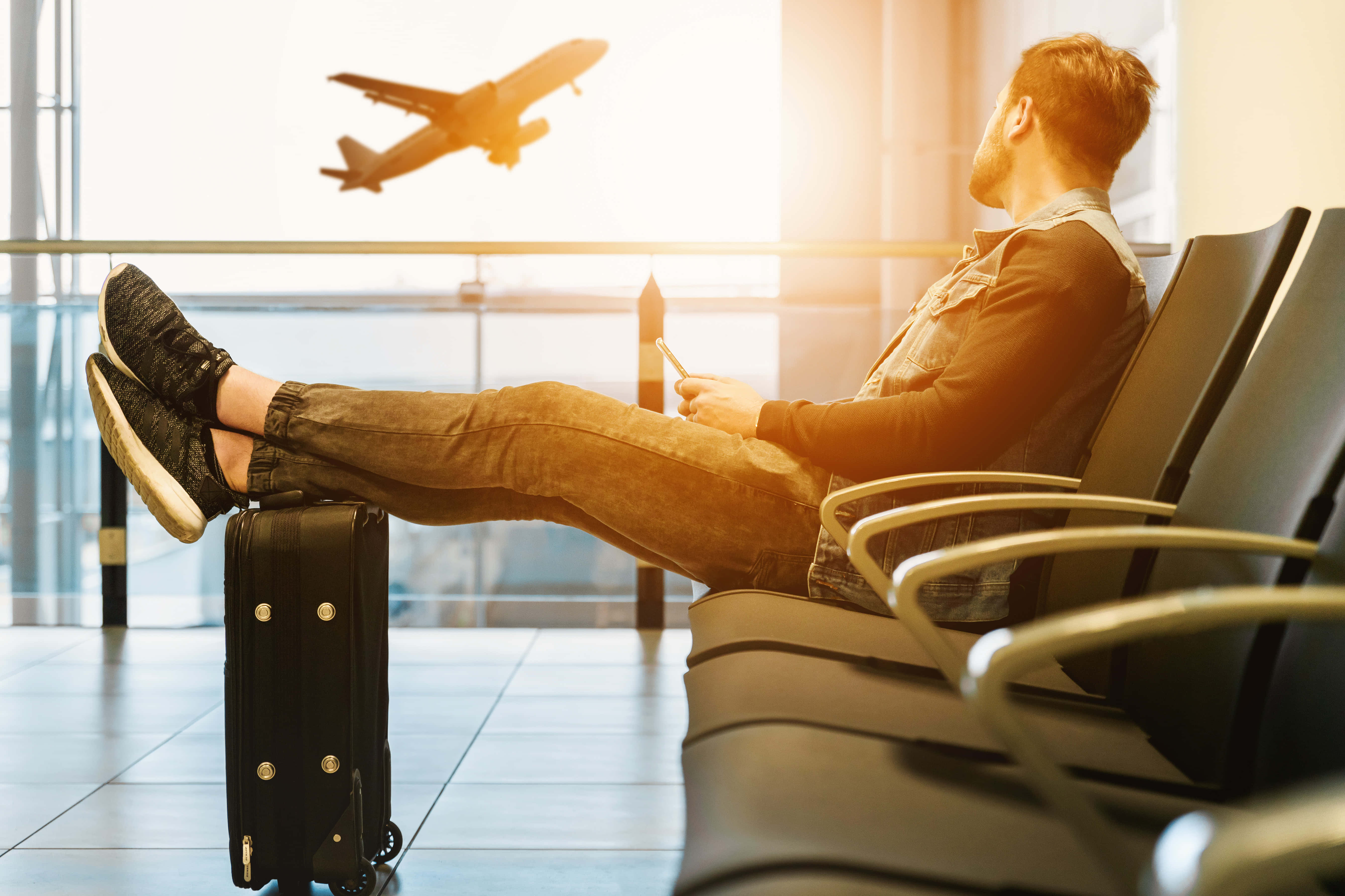 World Travel 桌布 壁紙 — 一名男子在機場航廈放鬆，雙腳擱在行李箱上，飛機在明亮的陽光窗戶外飛過。
