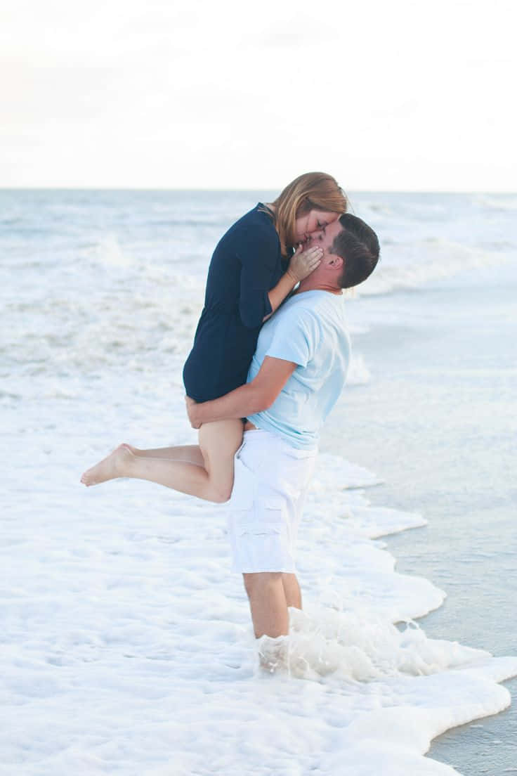 Kissing 桌布 壁紙 — 一名男子在海邊溫柔地抱起女子親吻，輕柔的海浪在他們腳邊翻滾。