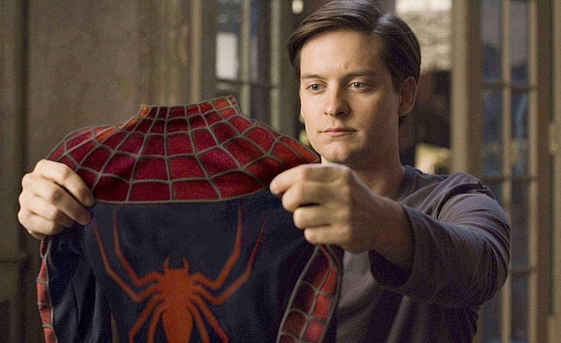 Spider Man 2 桌布 壁紙 — 一名男子在溫暖照明的室內環境中舉起一件帶有橙色蜘蛛標誌的紅藍色蜘蛛人戰衣。