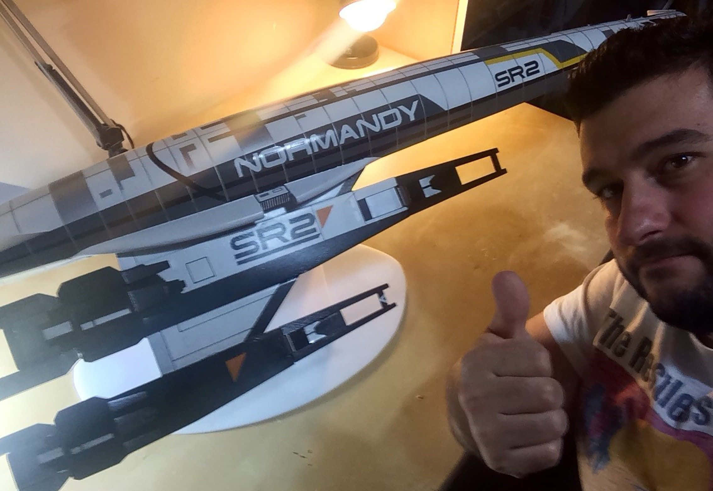 Normandy Sr-2 桌布 壁紙 — 一名男子在溫暖的室內燈光下，在詳細的Normandy SR2太空船模型旁邊豎起大拇指。