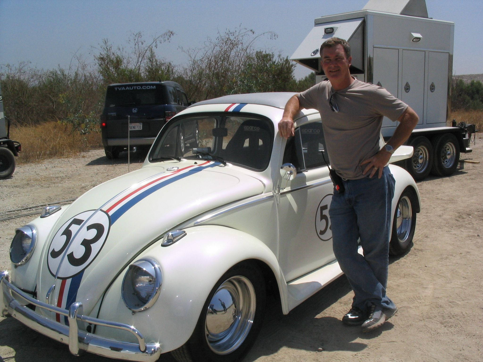 Herbie 桌布 壁紙 — 一名男子在白色復古賽車旁微笑，賽車帶有紅藍條紋和53號，停在塵土飛揚的地形上。