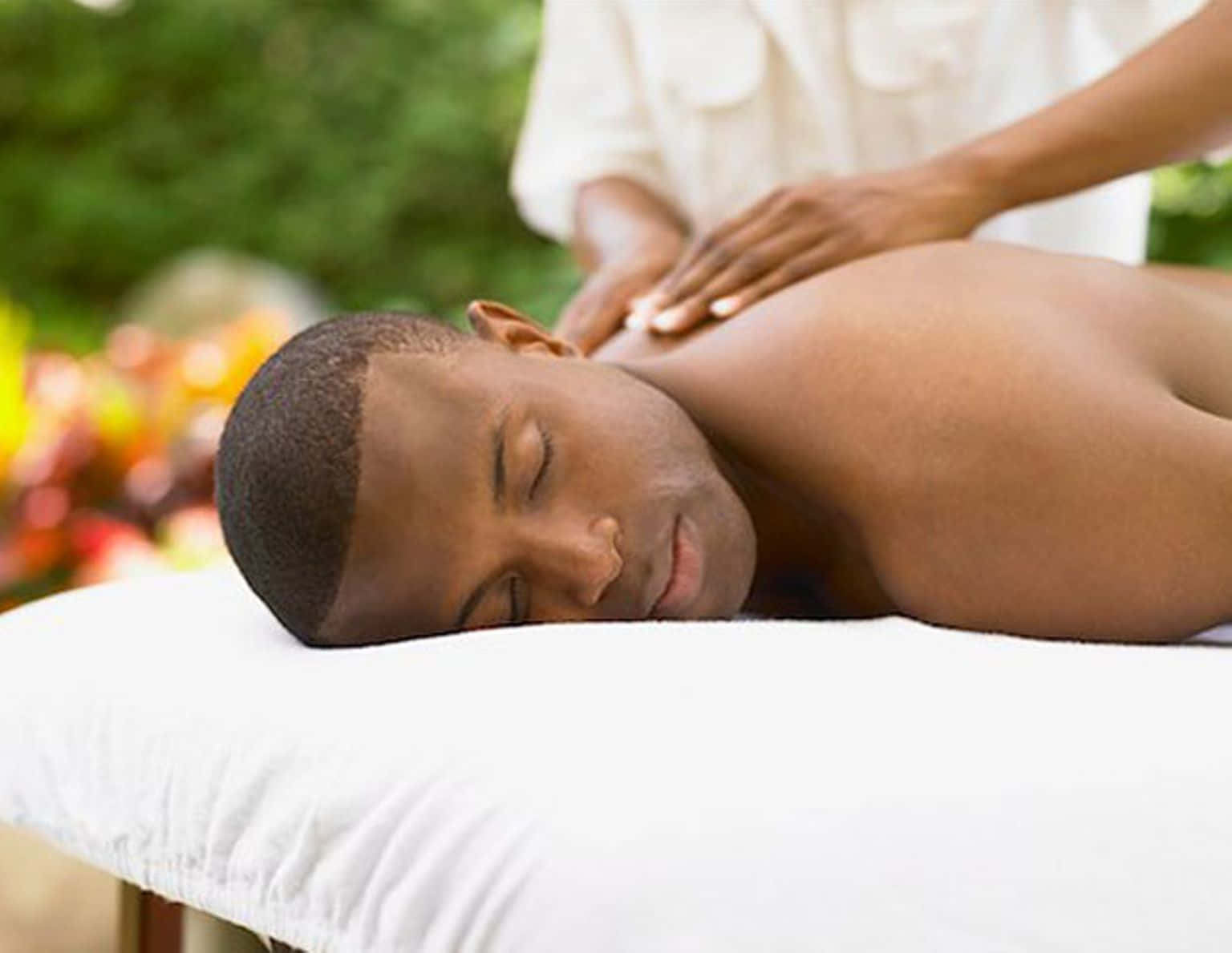 Massage 桌布 壁紙 — 一名男子在白色按摩床上平靜地休息，治療師在翠綠的戶外環境中輕柔地為他按摩肩膀。