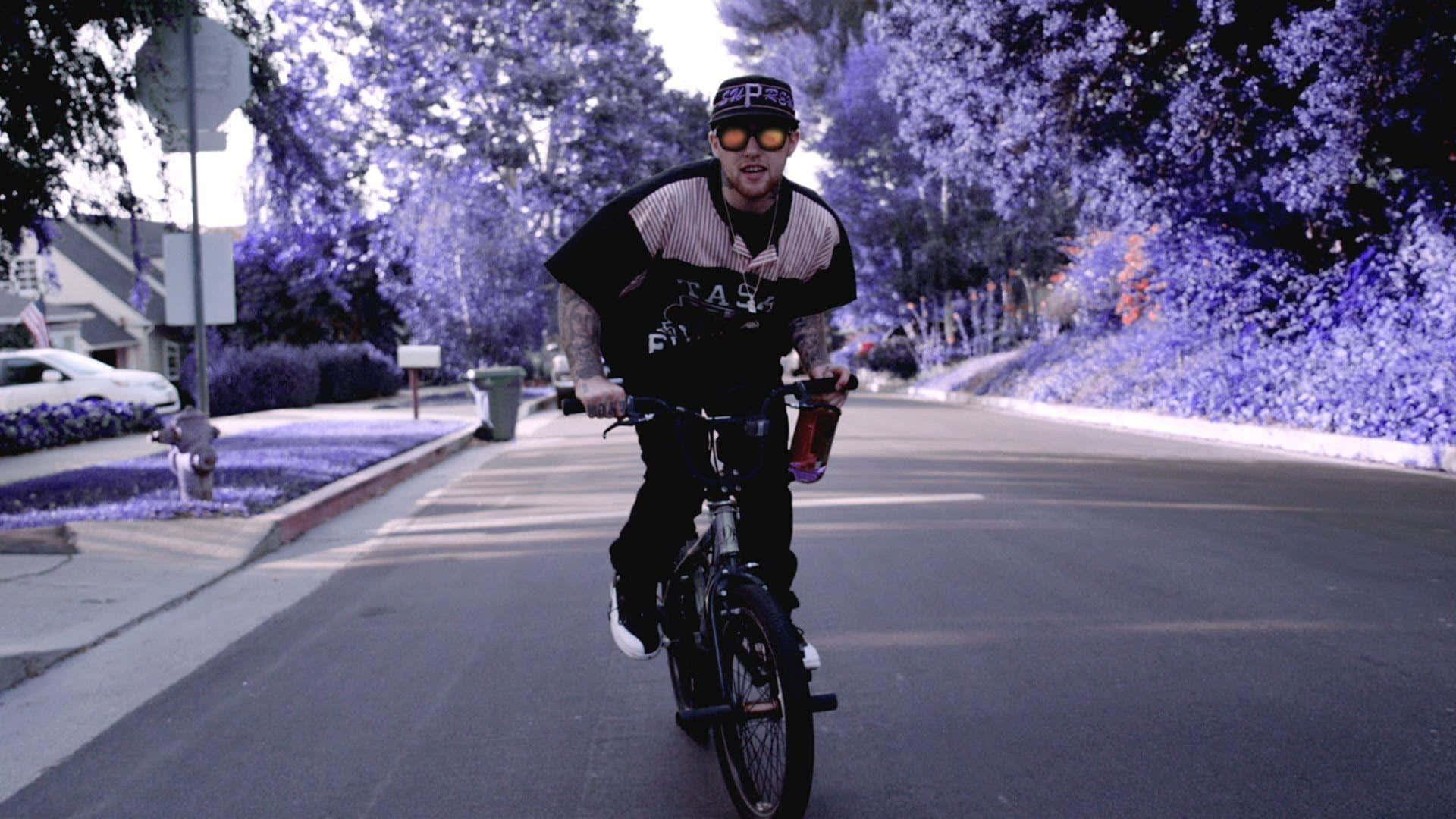 Mac Miller 桌布 壁紙 — 一名男子在超現實的紫色天空下騎腳踏車沿著郊區街道行駛，樹木以單色調發光。