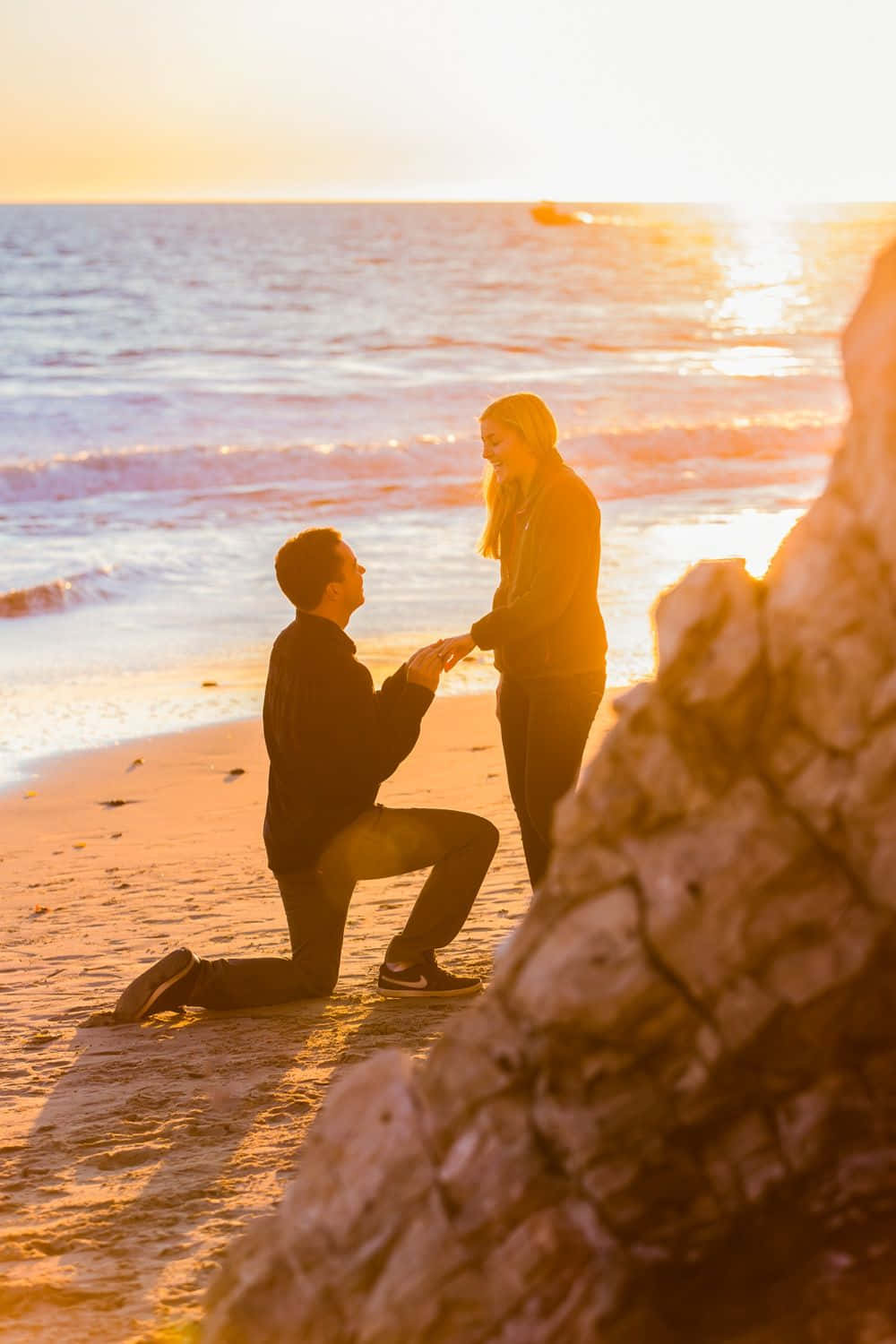Proposal 桌布 壁紙 — 一名男子在陽光明媚的海灘上單膝跪地，向一位微笑的女子求婚，金色的夕陽光芒灑滿海洋和岩石。