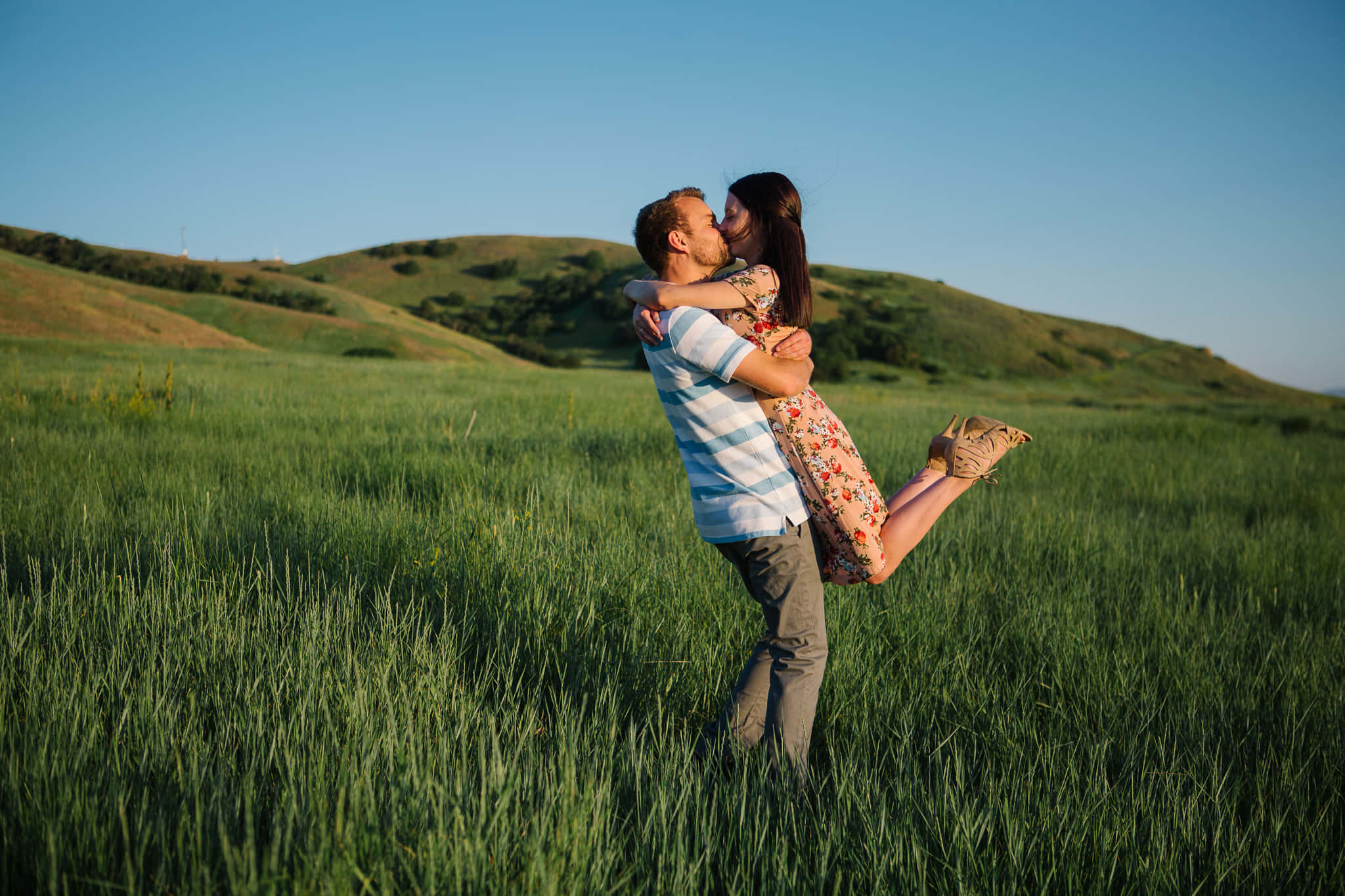 couple love 桌布 壁紙 — 一名男子在陽光明媚的綠色田野中，在晴朗的藍天下，將一名穿著花卉連衣裙的女子舉起親吻，背景是起伏的山丘。