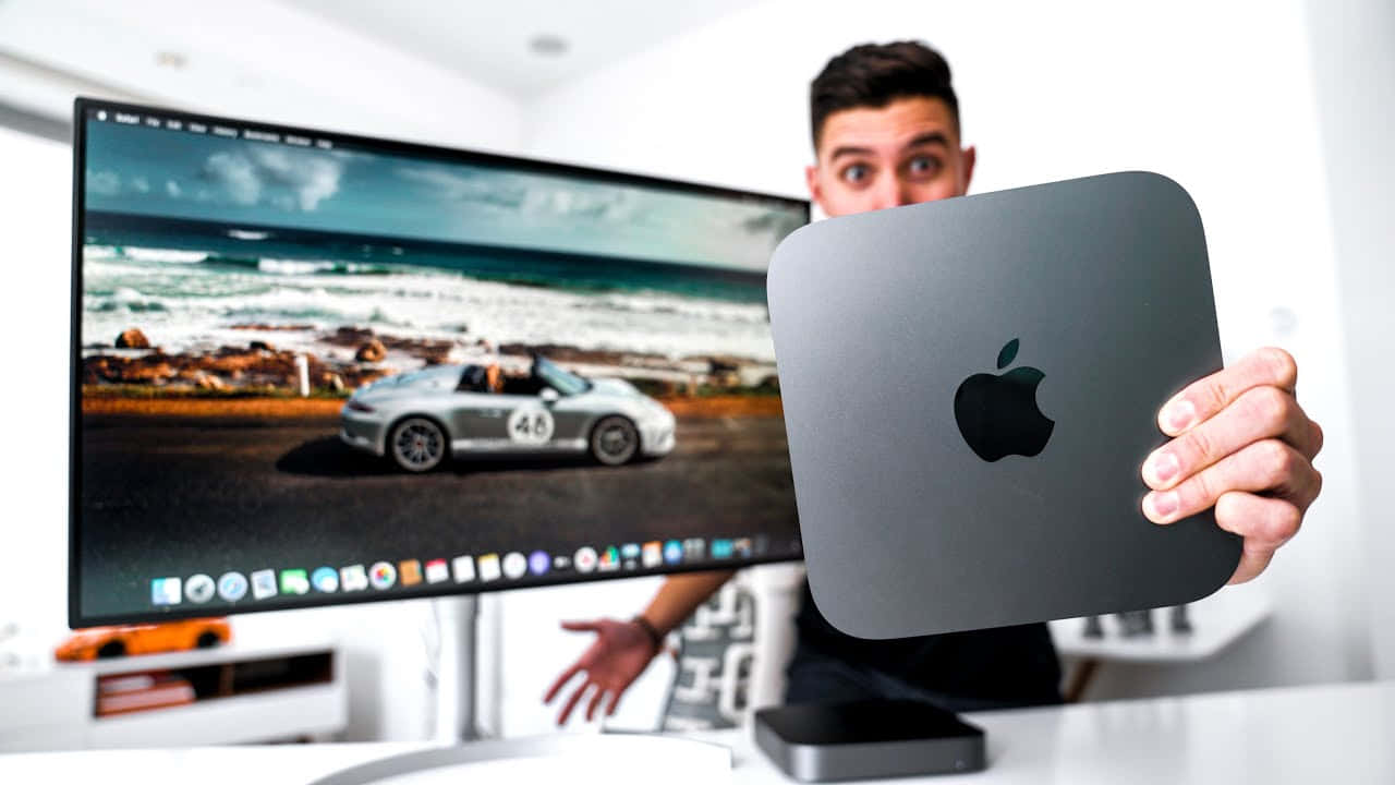 Mac 桌布 壁紙 — 一名男子在顯示器前舉起銀色 Mac mini，顯示器顯示沿海道路上的銀色賽車。