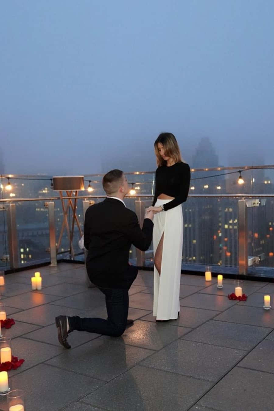 Proposal 桌布 壁紙 — 一名男子在黃昏時分跪在霧氣瀰漫的屋頂上，向一位身穿黑色上衣和白色開衩裙的女子求婚，周圍環繞著蠟燭和紅玫瑰。