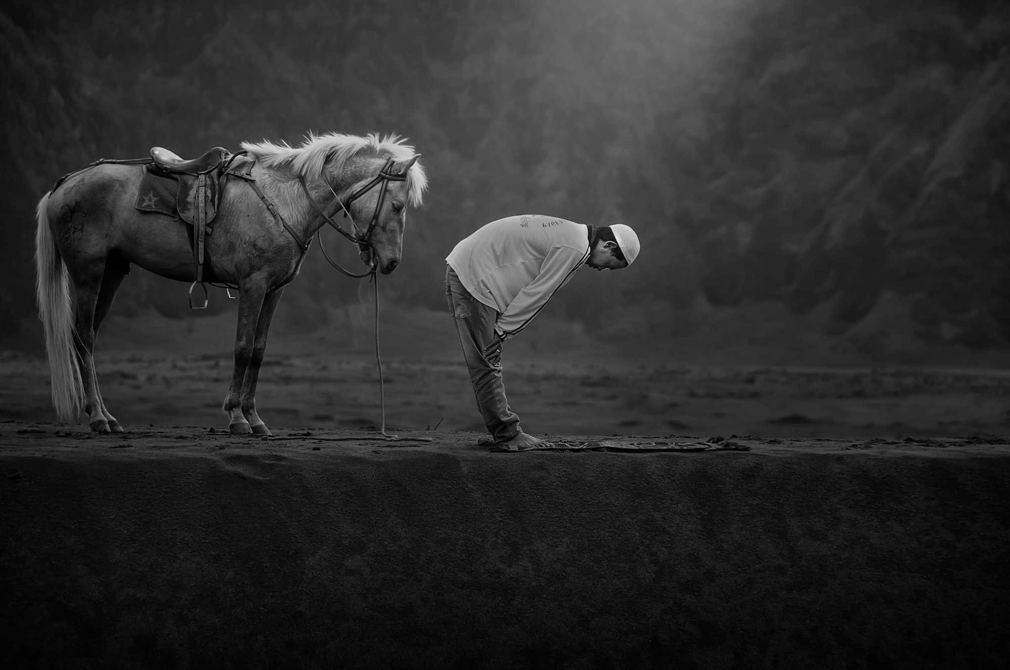 Praying 桌布 壁紙 — 一名男子在黎明時分，於霧氣瀰漫的山脊上，在他備好鞍的馬旁鞠躬祈禱，以單色呈現。