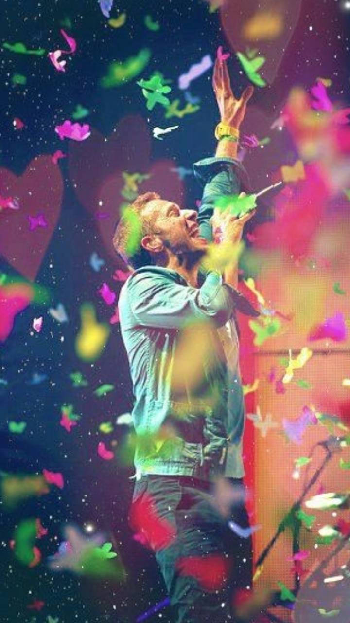 Chris Martin 桌布 壁紙 — 一名男子在黑暗舞台背景下，快樂地向上伸手，五彩紙屑和蝴蝶在他周圍旋轉。