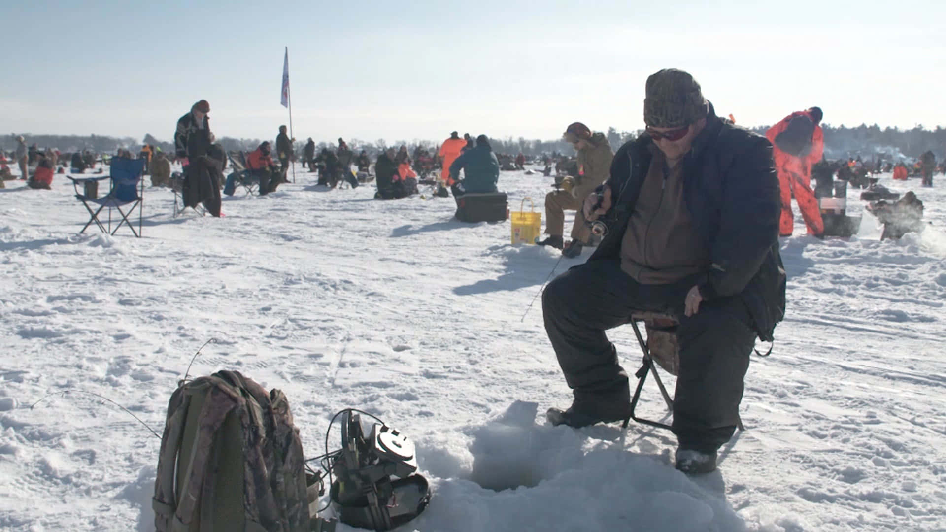 Ice Fishing 桌布 壁紙 — 一名男子坐在雪覆蓋的冰凍湖面上的凳子上，在晴朗的藍天下與其他人一起冰釣。