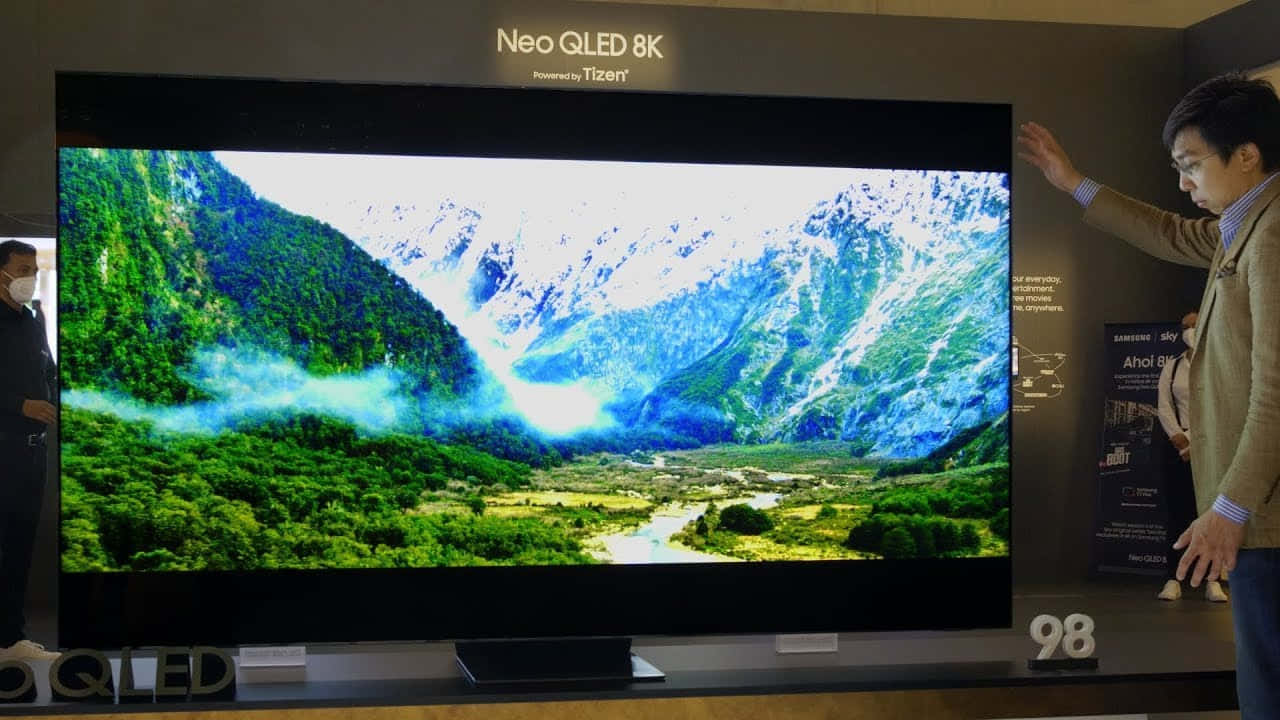 科技動圖 桌布 壁紙 — 一名男子對著一台巨大的98吋Neo QLED 8K電視比手勢，電視上顯示著生動的山谷景色，有白雪覆蓋的山峰和茂密的綠色森林。